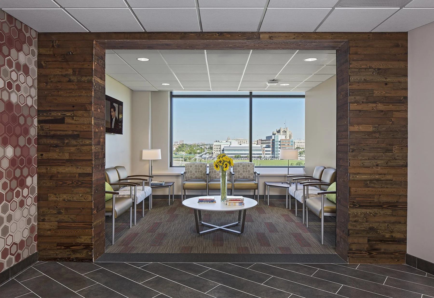 Office waiting area with beige chairs, a white round table with books and a vase of sunflowers, large window with city view, wood-paneled walls, and patterned wallpaper on the left.