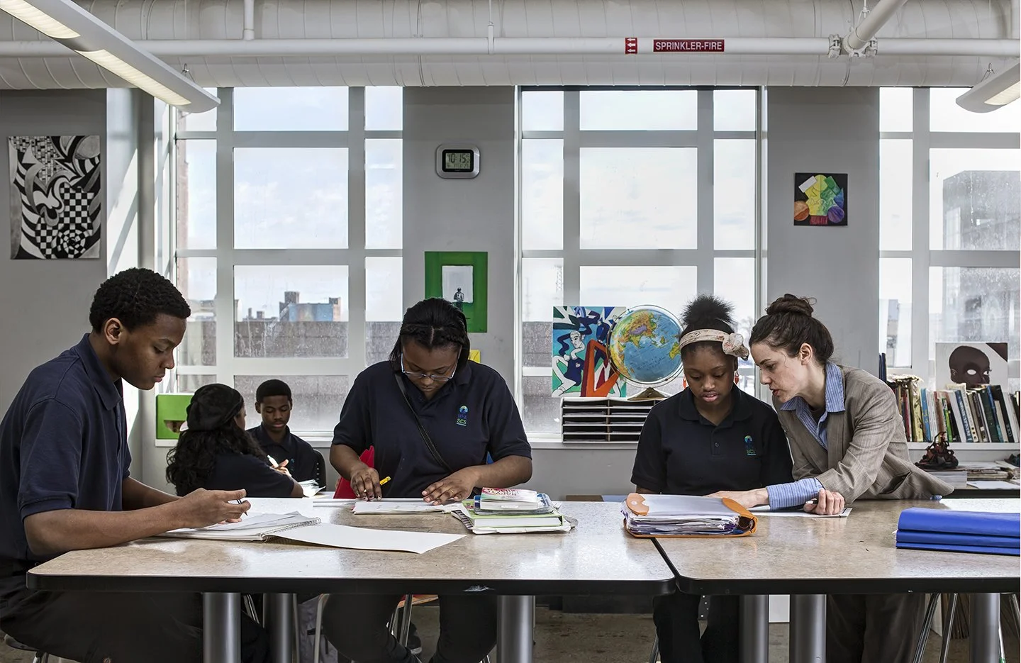 A teacher and students working on assignments at a classroom table with large windows in the background.