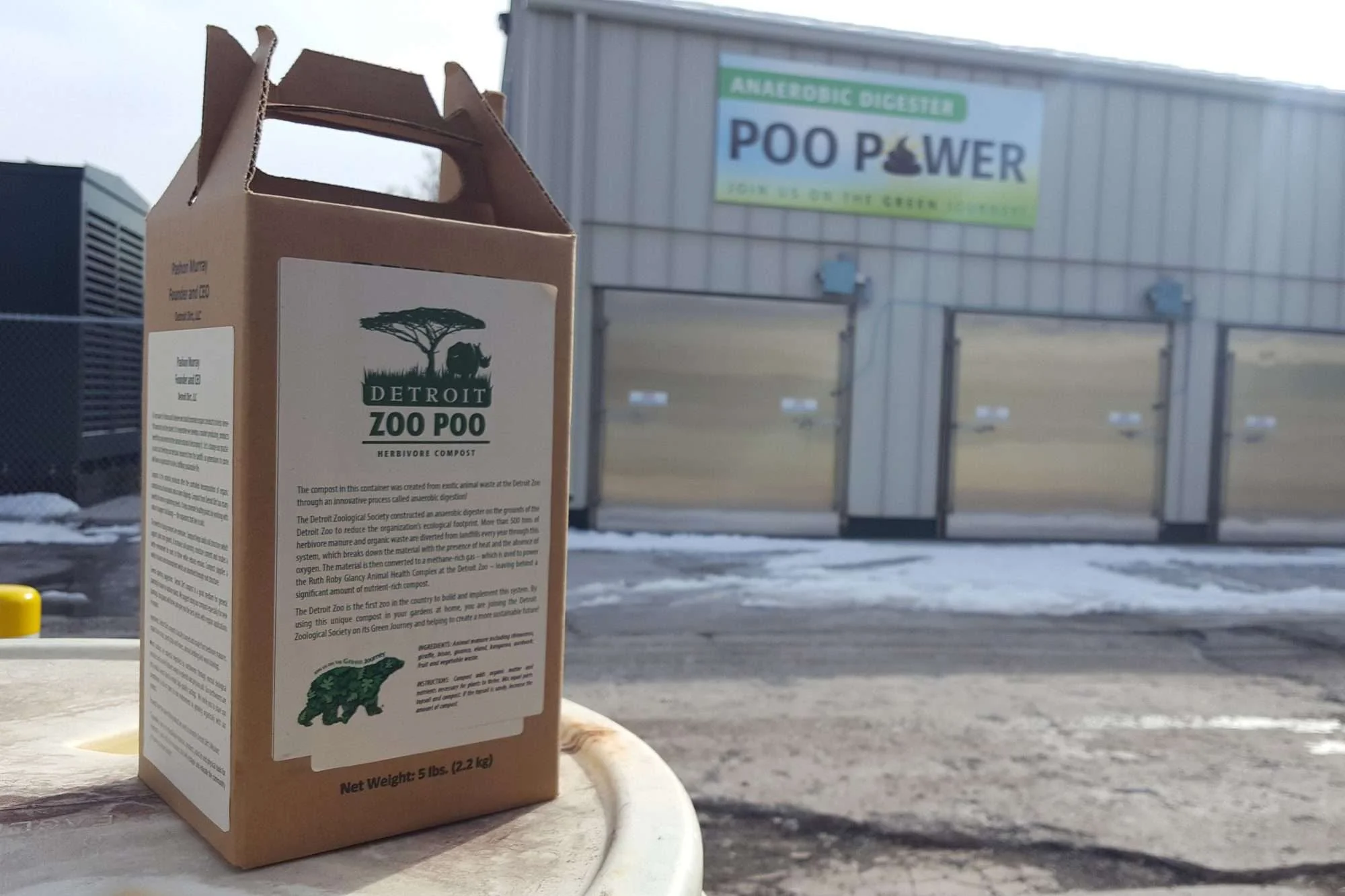 A small cardboard box labeled 'Detroit Zoo Poo' on a white surface outdoors, with a sign for an anaerobic digester named 'Poo Power' in the background.