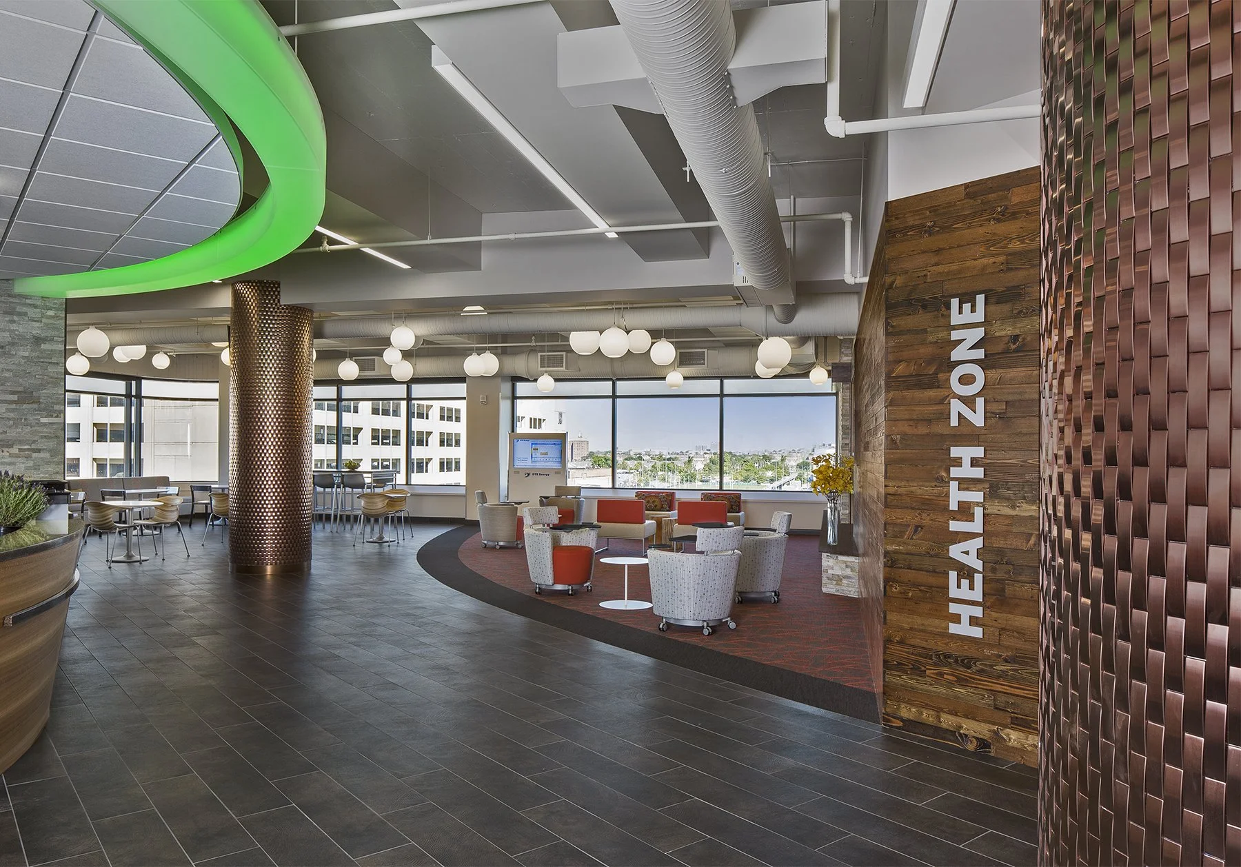 Interior view of a modern healthcare facility or office lounge area with seating, large windows, decorative lighting, and a sign that reads 'HEALTH ZONE' on a wooden wall.