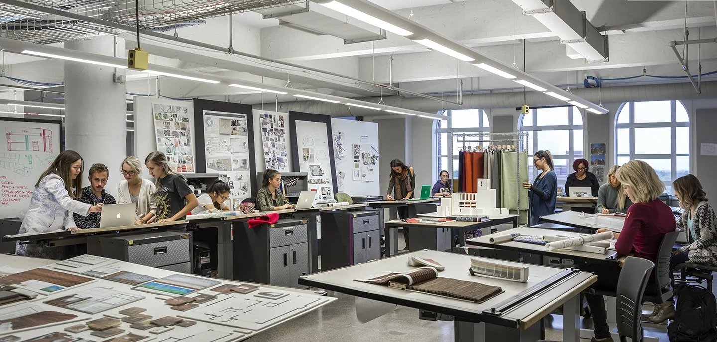 A classroom or studio with multiple women working on laptops, documents, and design projects, with large windows and posters on the walls.