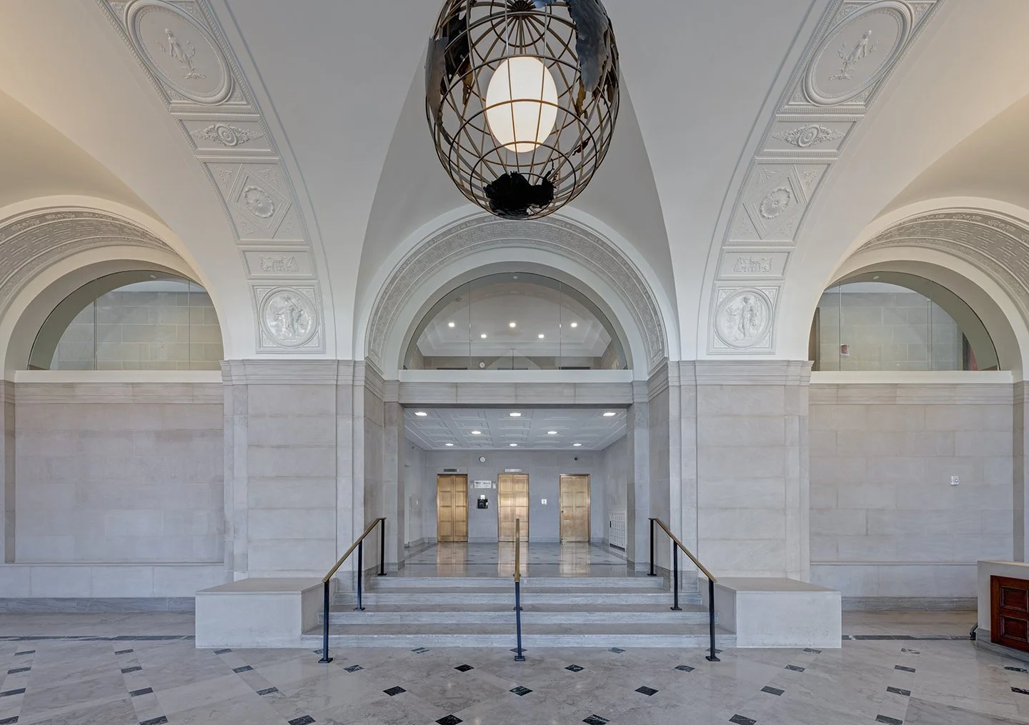 Empty lobby with marble floors, arched doorways, high ceilings, and a large hanging light fixture. There are two elevators at the back with closed doors.