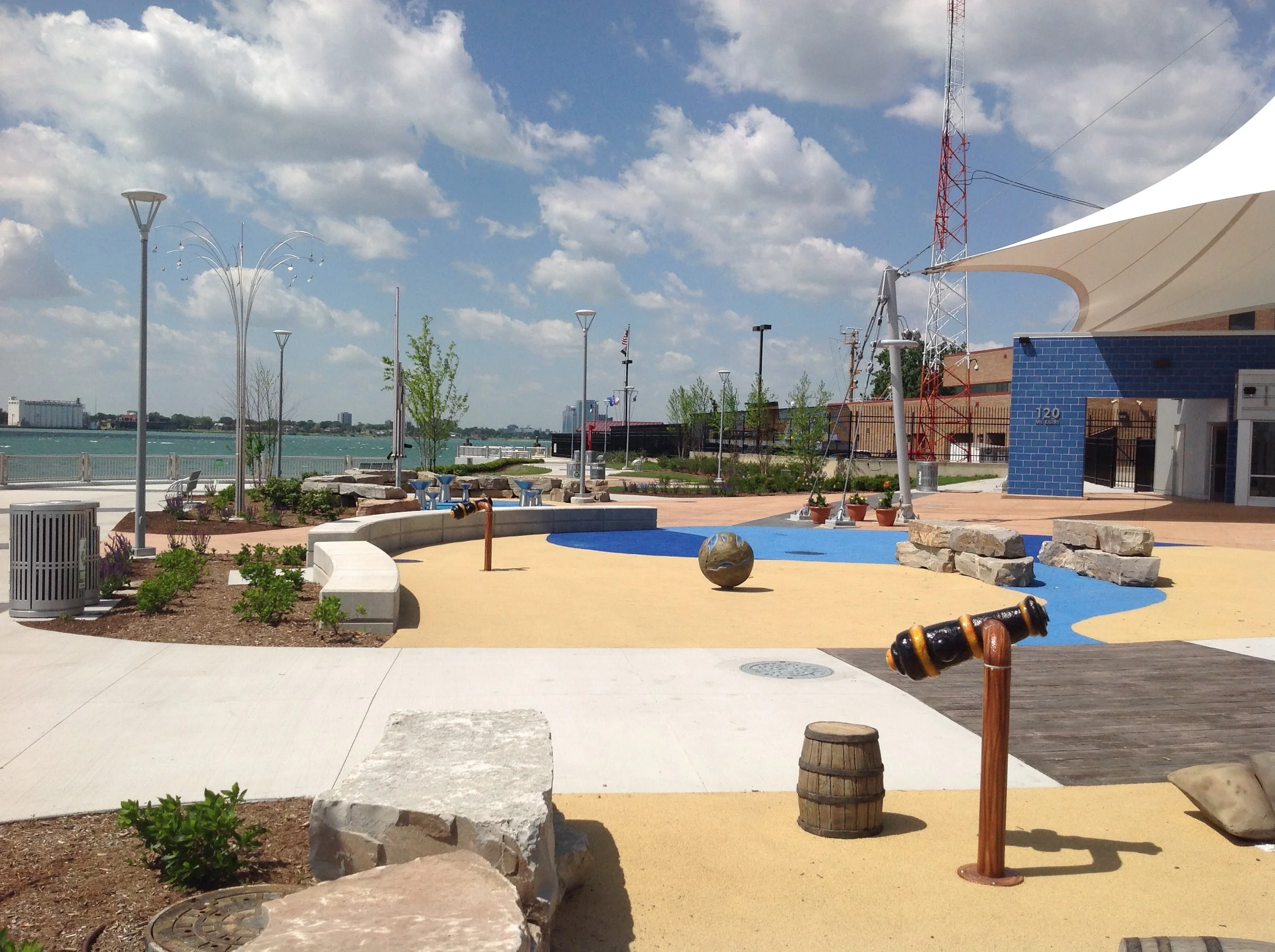 A park area by a body of water with a blue sky and scattered clouds. There are several street lamps, small trees, a fountain, and a mid-sized building with a blue tiled wall and a white canopy. A red and white radio tower is visible in the background