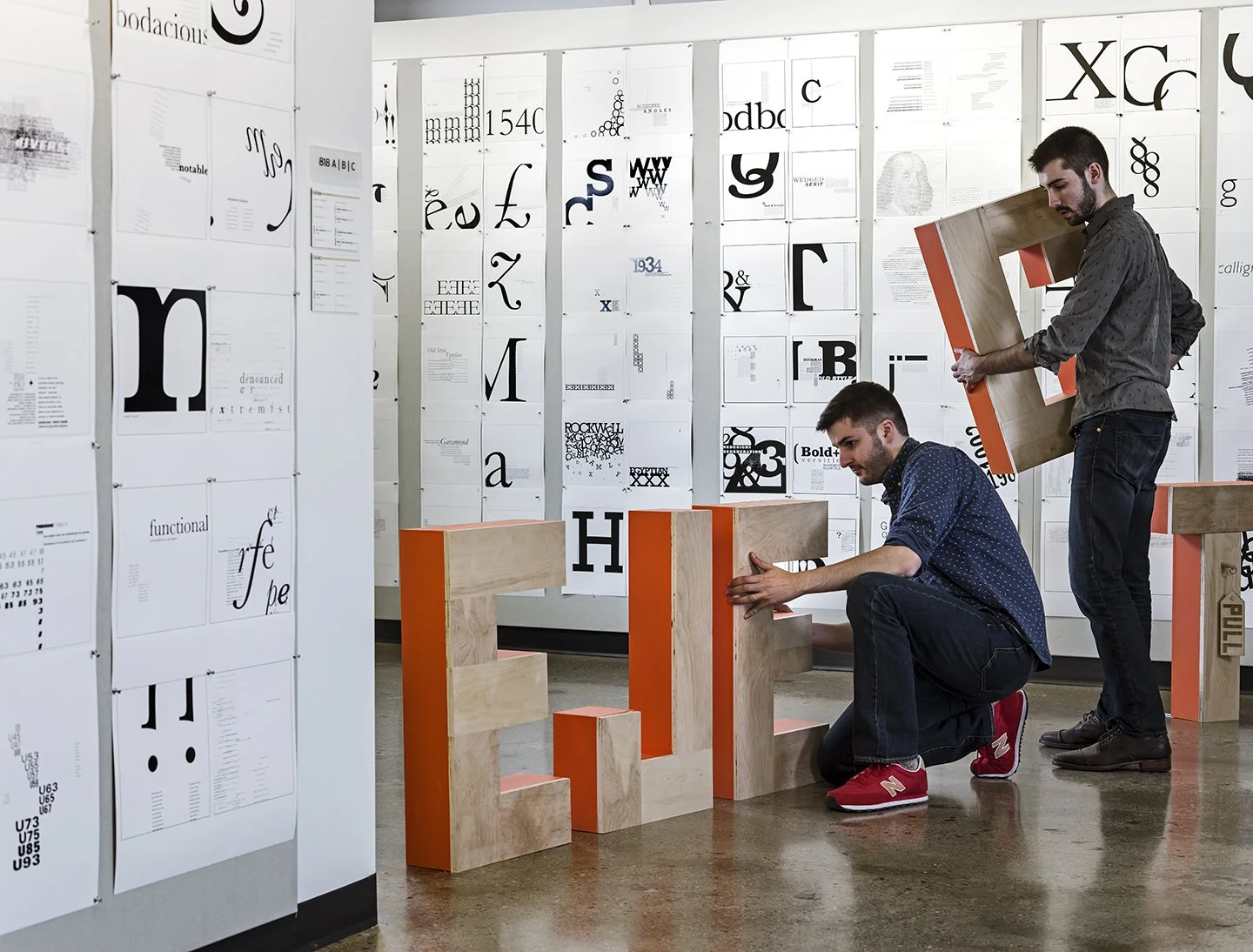 Two men assembling large wooden letter sculptures in an art gallery with typeface posters on the walls.