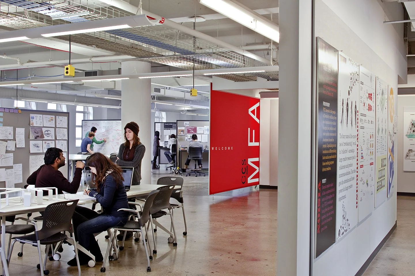 A modern office space with people working and collaborating at tables and desks, decorated with posters and artwork on the walls, and a large red banner with white text welcoming to a conference area.
