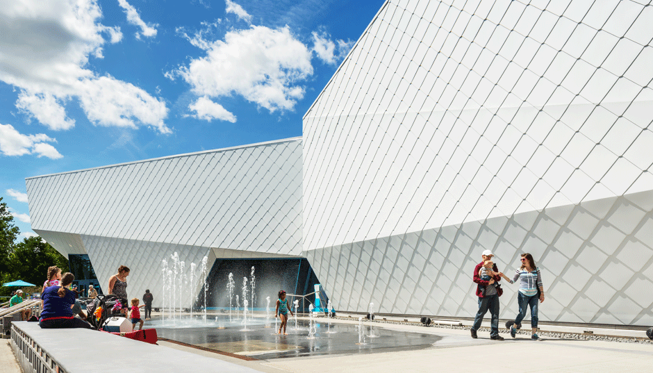 People enjoying a water fountain outside a modern, geometric white building on a sunny day.