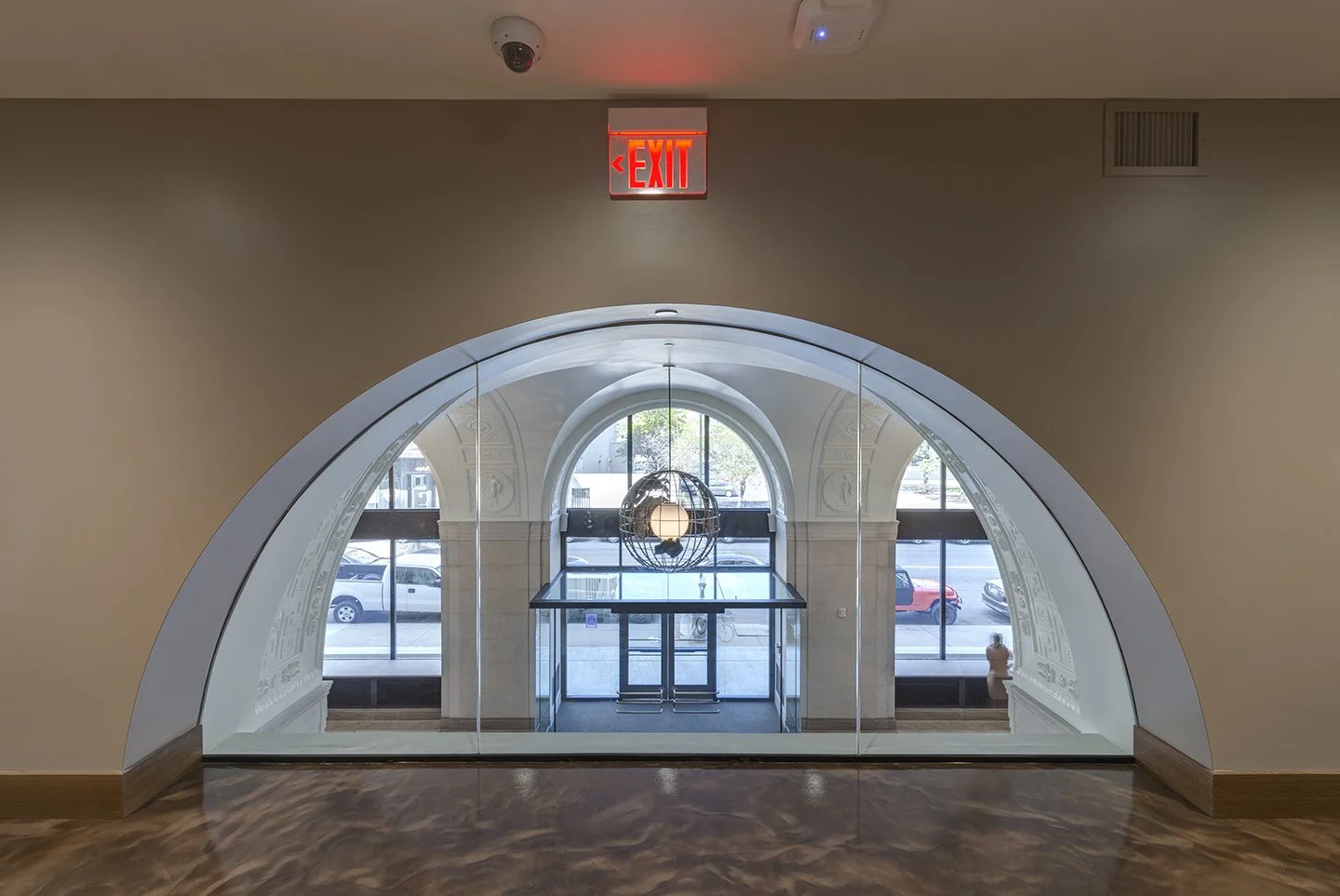 Interior view through a glass door with an archway, showing a lobby area with a window, a hanging spherical chandelier, and parked cars outside. An illuminated red 'EXIT' sign is above the door.