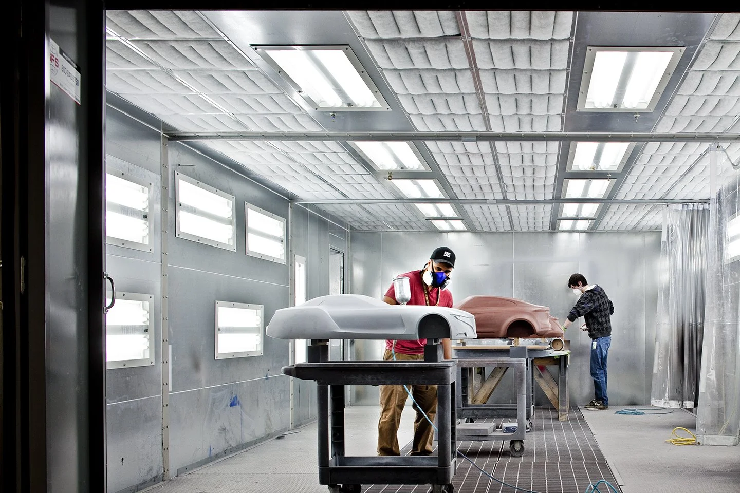Two workers in a spray booth working on car body parts, with one sanding a brown car panel and the other working on a white panel.