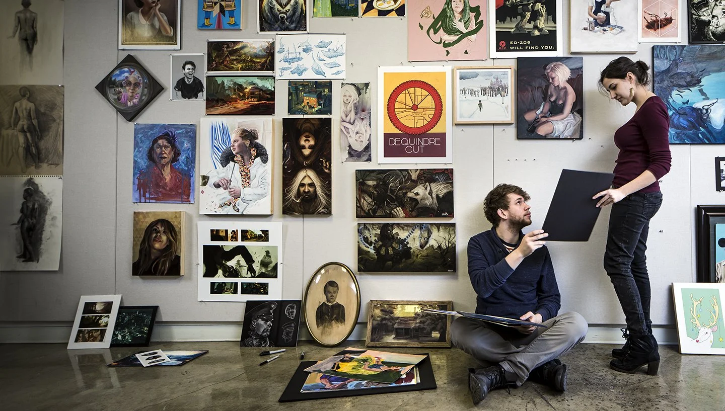 Two people in an art gallery, one sitting on the floor with a sketchpad and the other standing and holding a framed artwork, surrounded by various paintings and photographs on the wall and on the floor.
