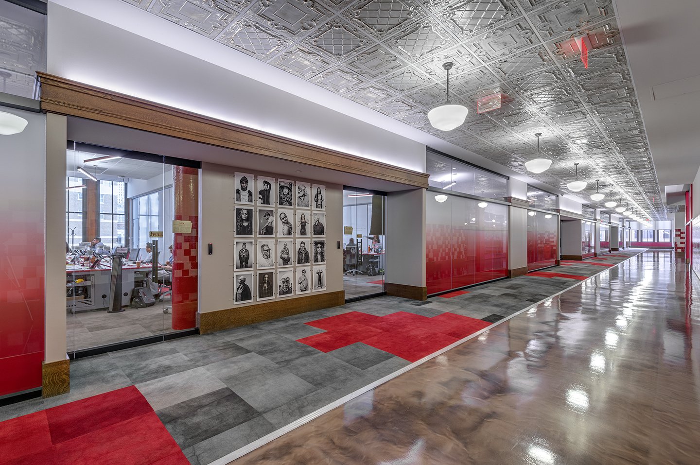 Office corridor with glass-enclosed meeting rooms, black-and-white photos on the wall, red and gray carpet tiles, and a shiny wooden floor.