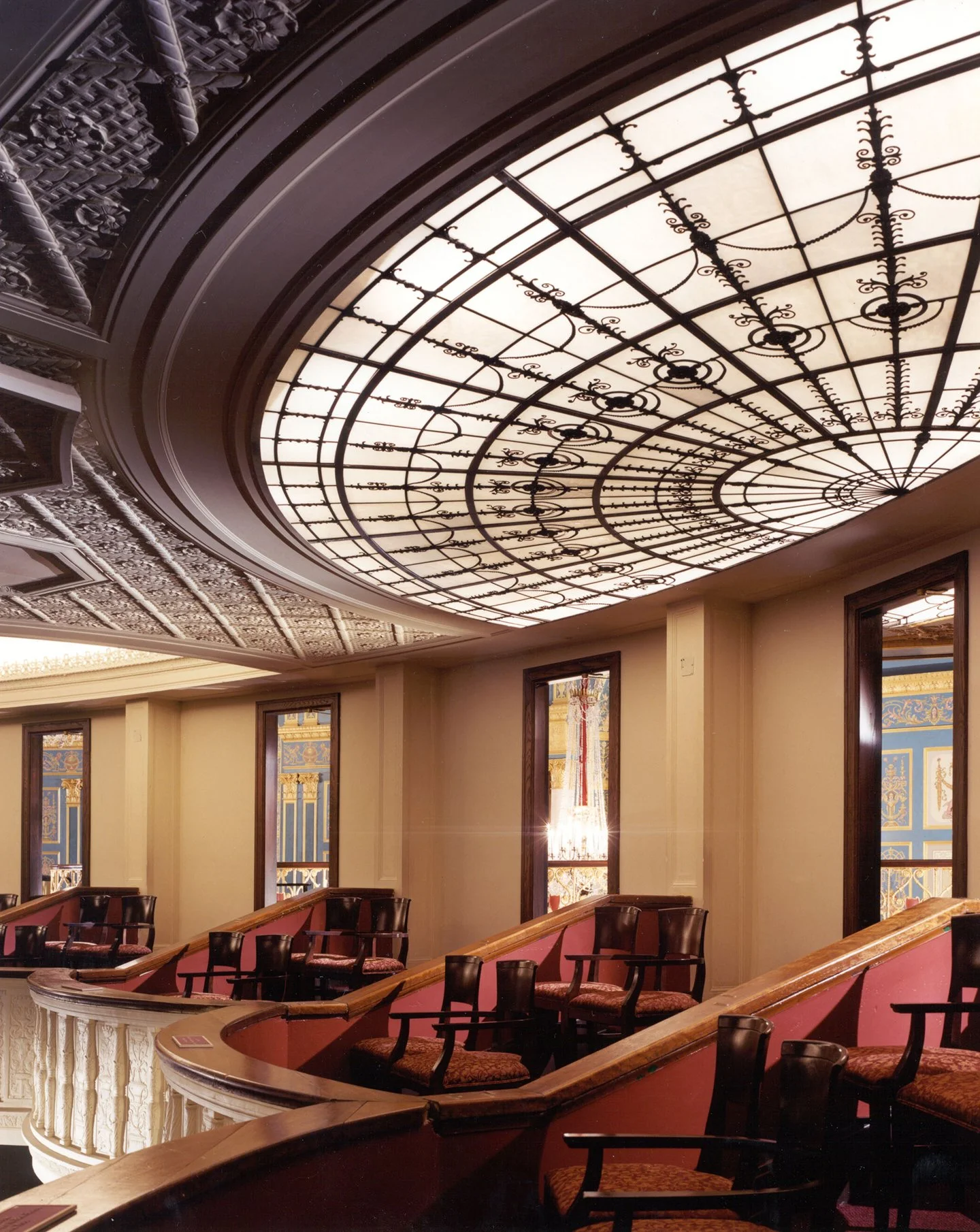 Interior of a theater with a decorative wrought iron ceiling and red velvet chairs arranged in ascending rows near the wall, with framed ornate wallpaper and large windows.
