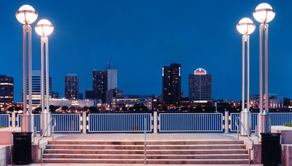 Nighttime city skyline viewed from a waterfront promenade with street lamps, steps, and metal railings.