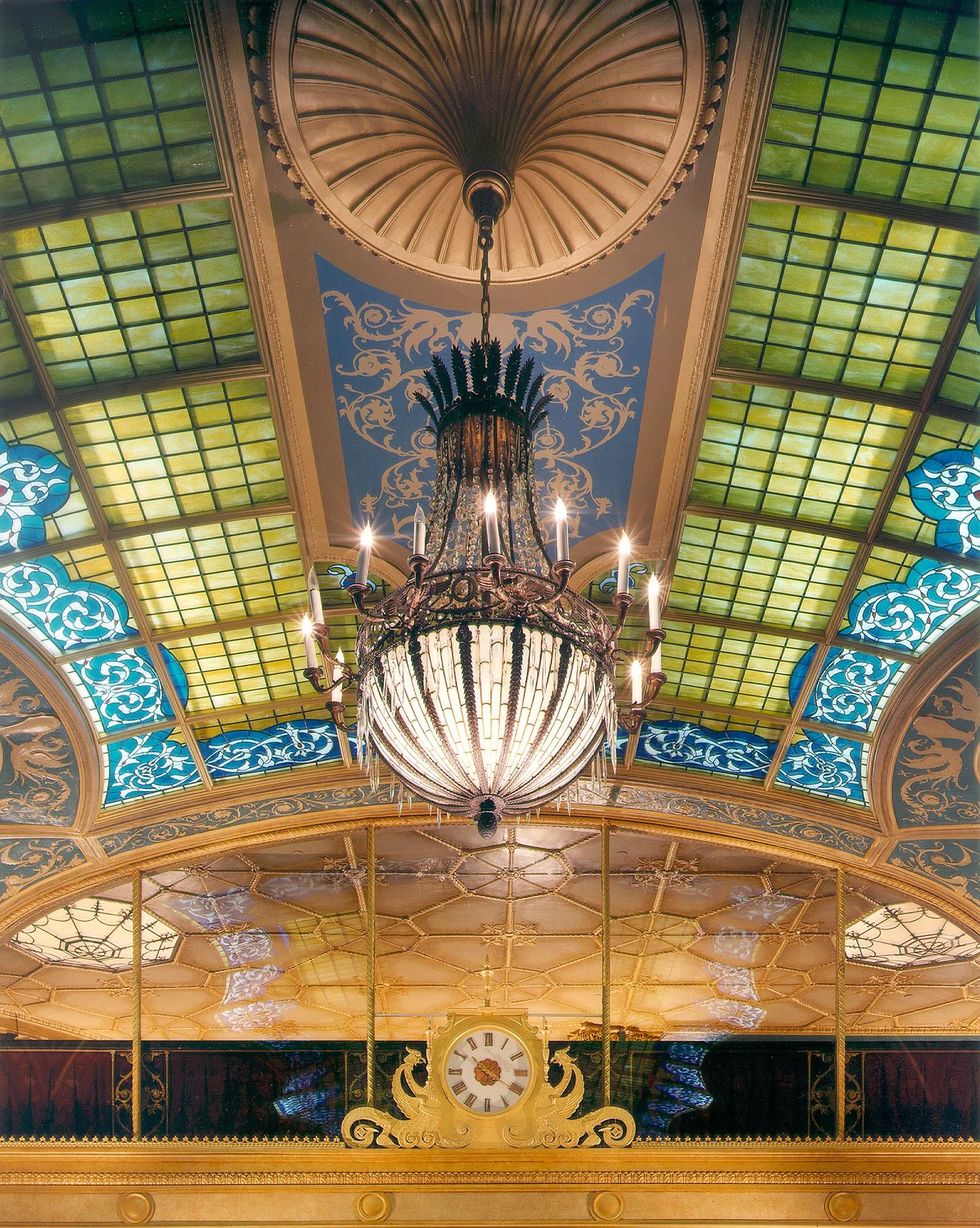 Ornate chandelier hanging from a detailed ceiling with stained glass panels, decorative patterns, and a clock below.