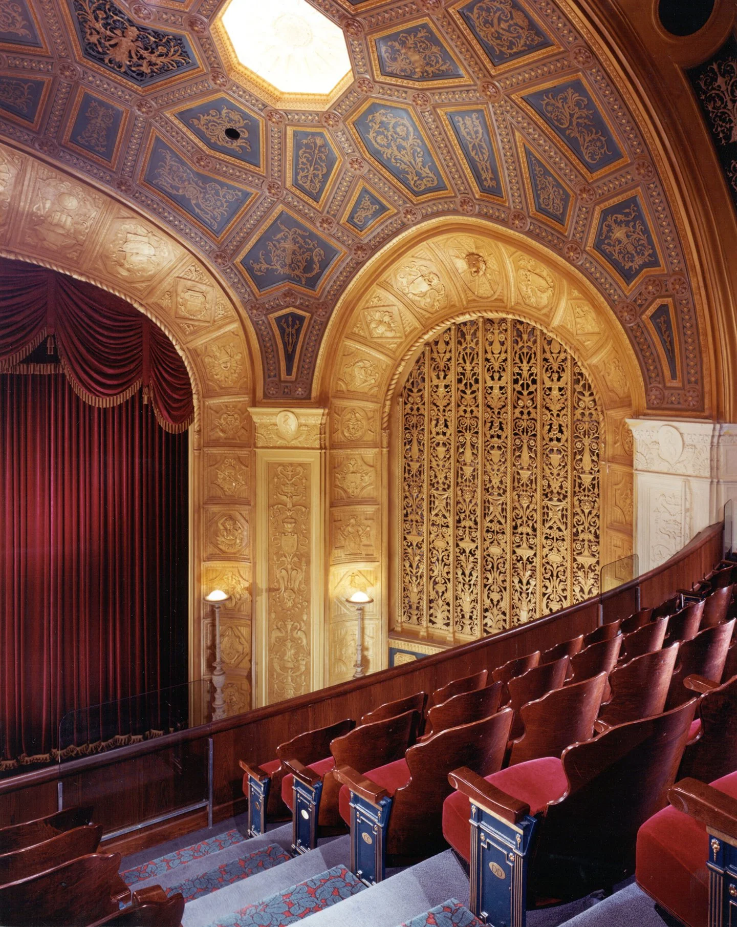 Interior view of a theater with ornate, gold decorative archways, blue and gold painted ceiling panels with intricate designs, red velvet curtains, wooden seats with red velvet cushions, and a staircase leading down to the stage area.