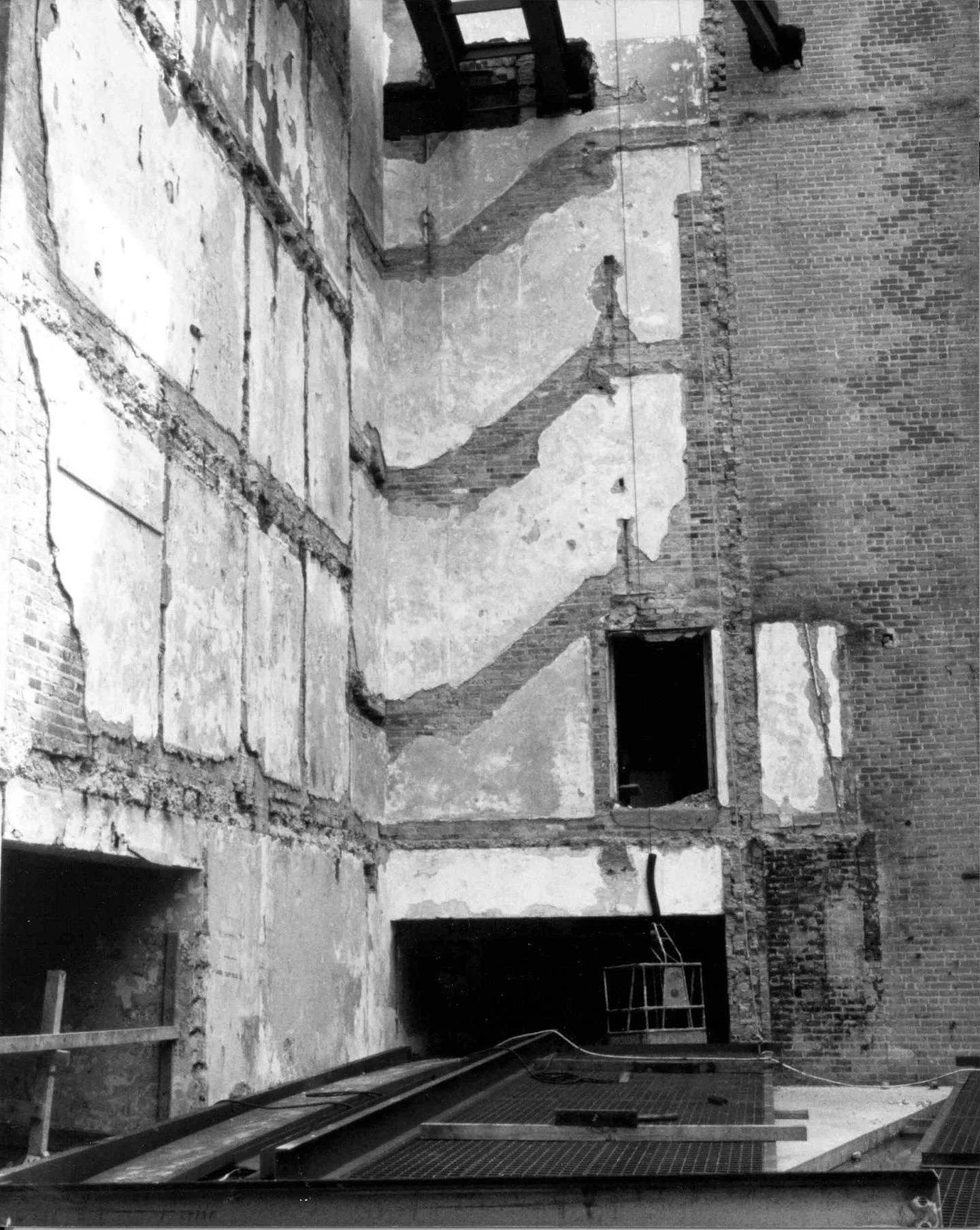 Black and white photo of a building interior under renovation, with exposed brick walls, a partially removed floor, and construction materials on the ground.