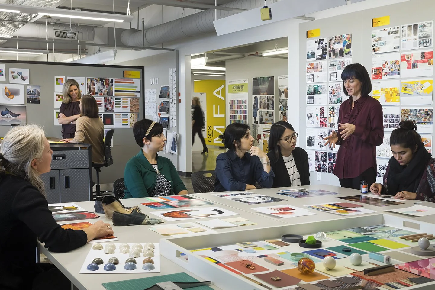 A woman in a burgundy shirt gives a presentation to a group of five women seated at a table in a design studio filled with colorful artwork and design material.