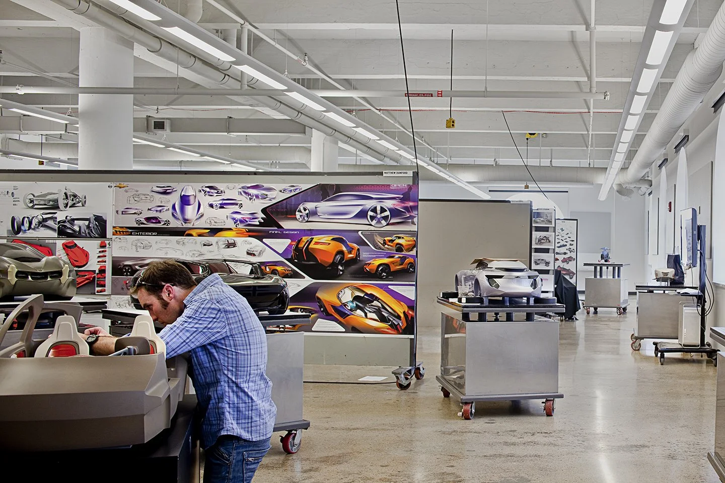 An industrial design studio with a man working on a model car. The background features concept art and designs for futuristic and sports cars displayed on large boards.