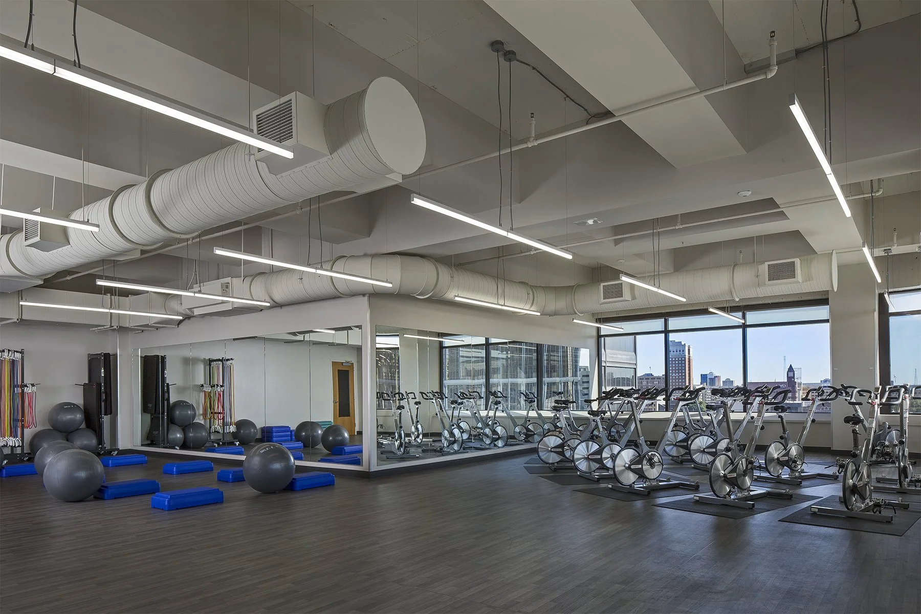Indoor gym with stationary bikes, elliptical machines, large exercise balls, resistance bands, mats, and weights. Bright space with large windows showing city buildings.