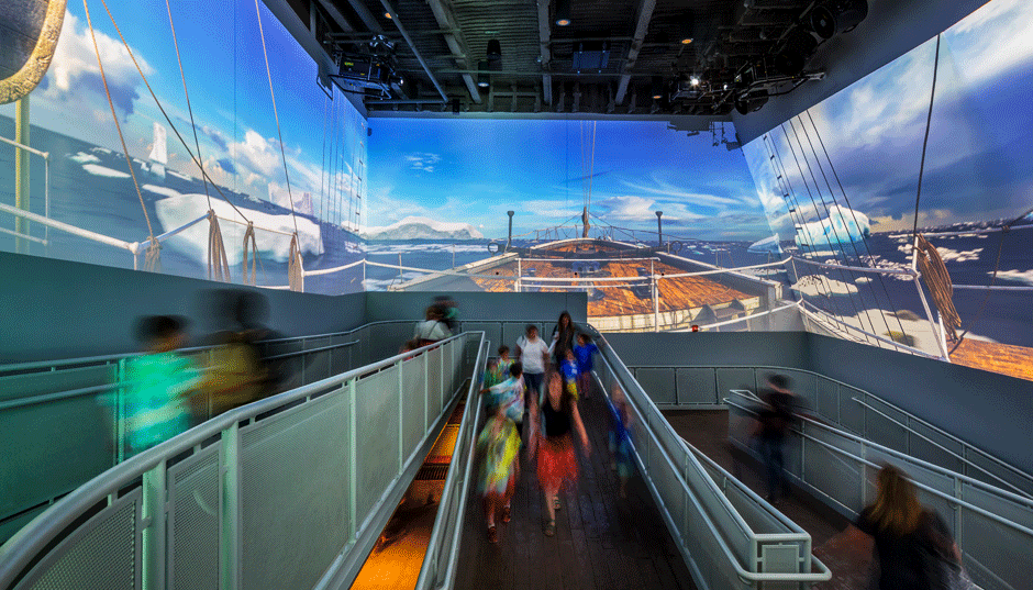 People walking on a curved indoor observation deck with panoramic digital sea and sky projections.