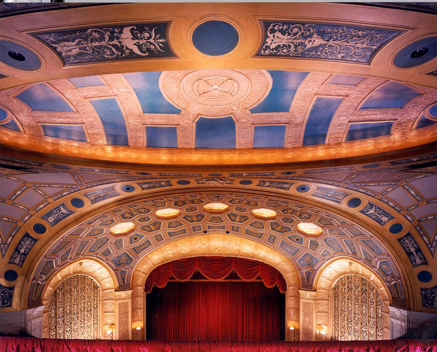 Interior view of a grand theater or opera house with an ornate, decorated ceiling, a stage with red velvet curtains, and seating area in front.