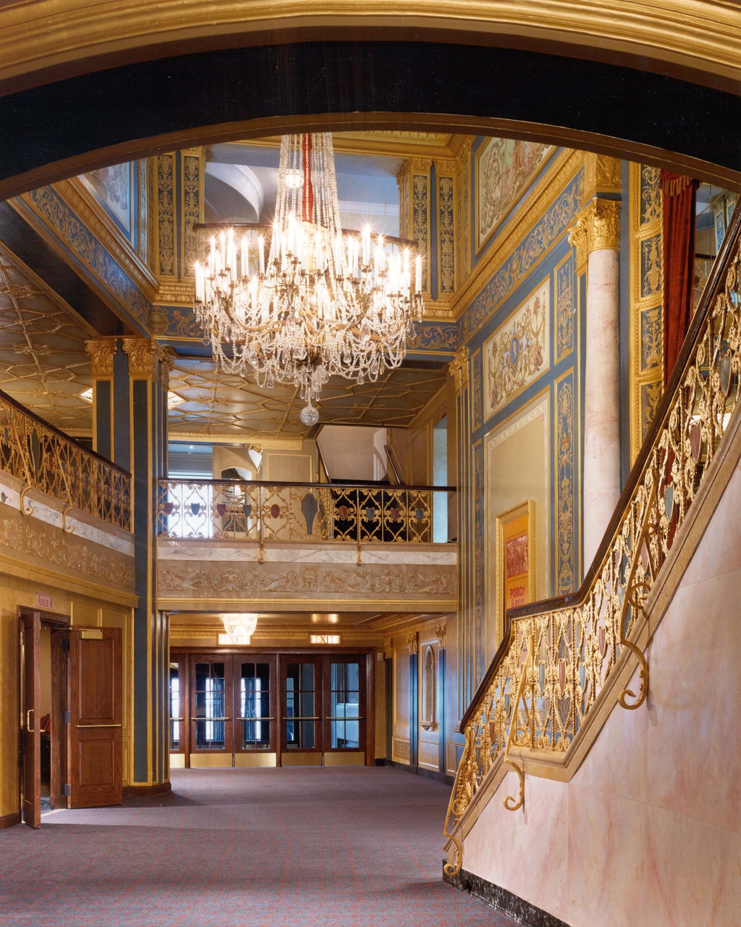 Elegant hotel lobby with ornate gold and blue decorative molding, a large crystal chandelier hanging from the ceiling, and a decorative staircase with gold railing on the right. Double doors are visible at the far end of the room.