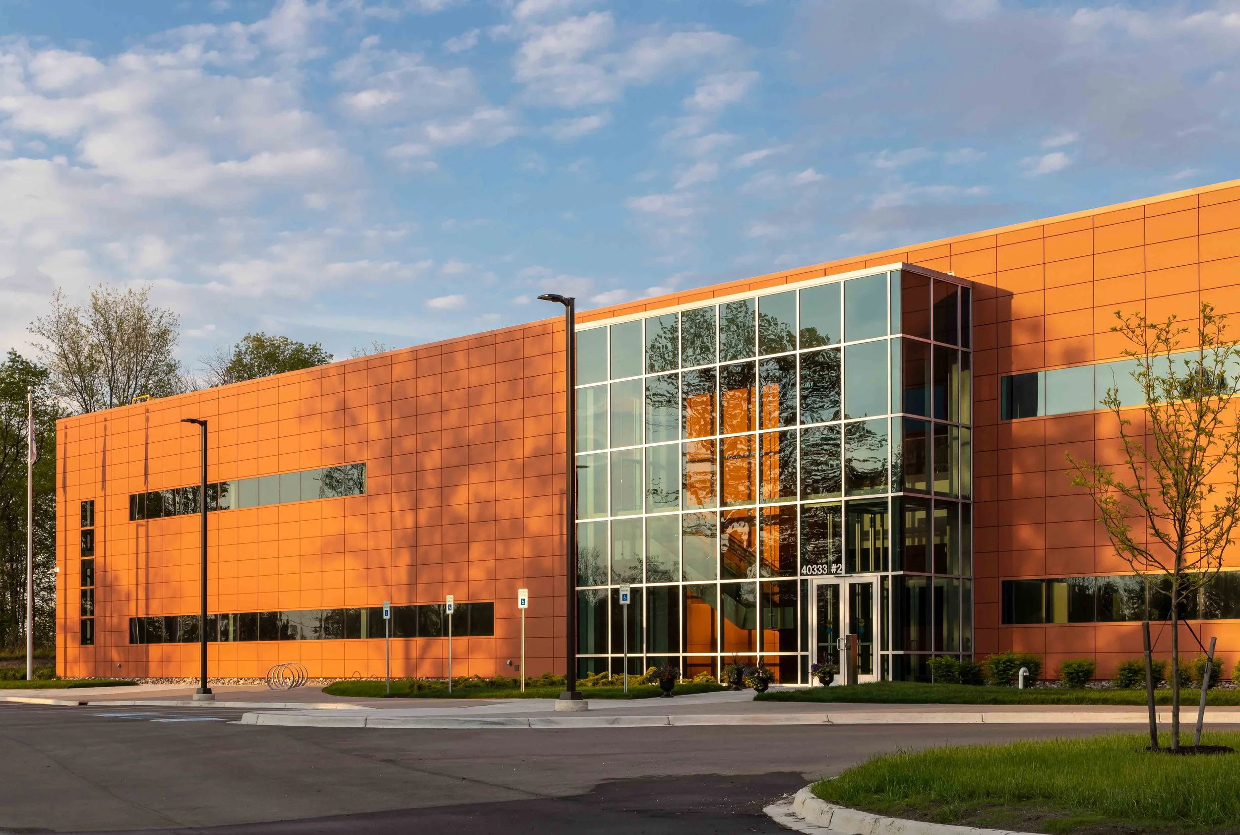 Modern building with orange panels and large glass windows, surrounded by trees and a parking lot with street lamps, under a partly cloudy sky.