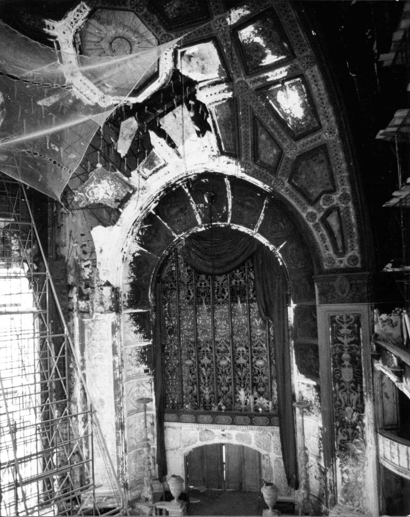 Black and white photo of a grand, ornate interior under renovation, featuring a large archway with intricate decorative details, scaffolding on the left side, and construction materials on the floor.