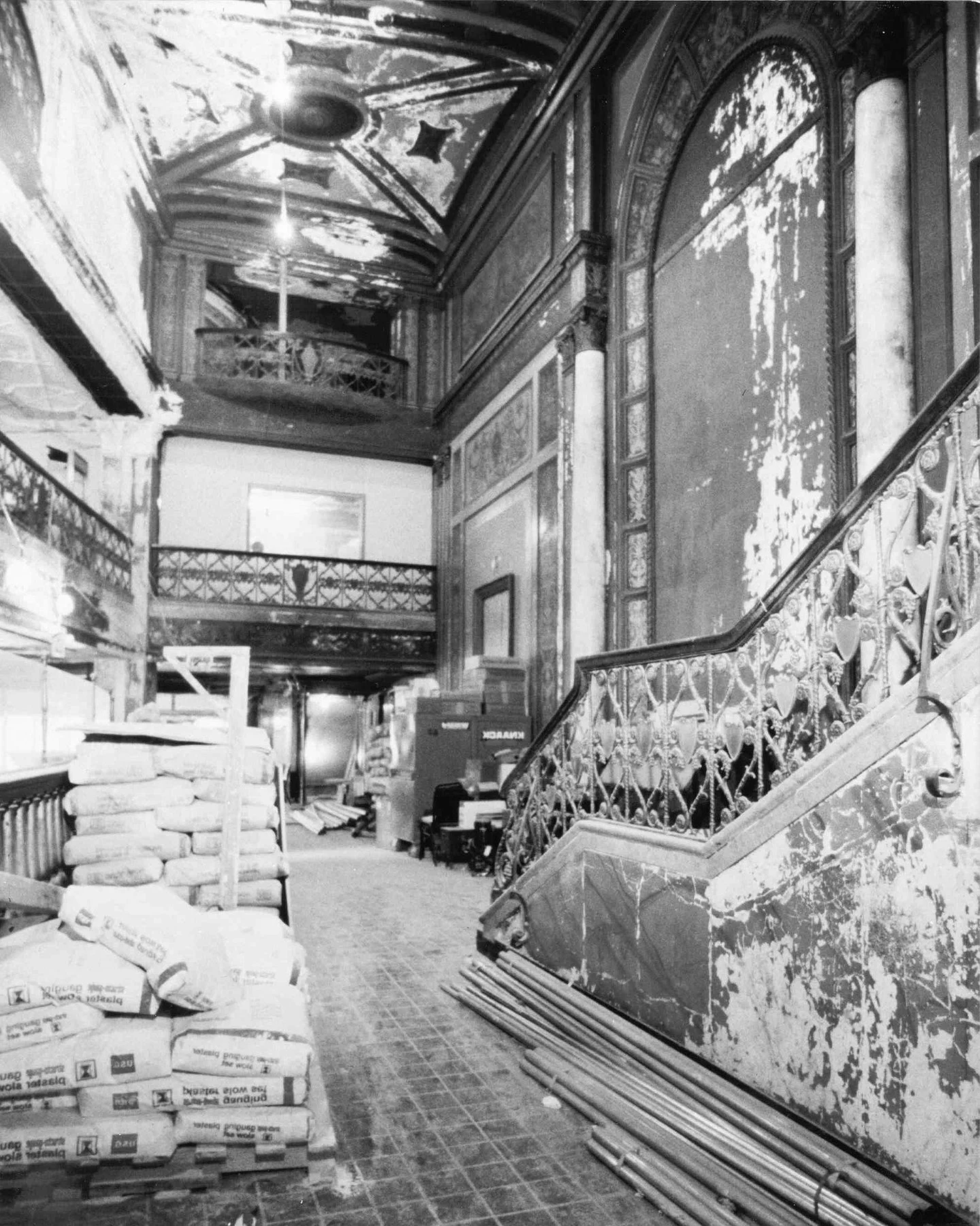Indoor construction site with bags of mortar, stacked wood planks, metal pipes, a metal staircase with ornate railing, and an ornate wall with peeling paint. The ceiling has decorative moldings and a chandelier.
