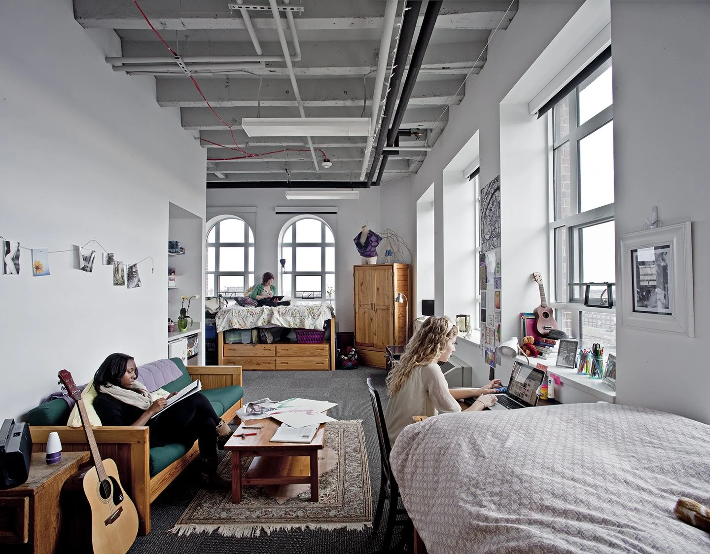 A spacious loft apartment with three women engaging in various activities: one reading on a couch, another working on a laptop at a desk, and the third sitting on a bed looking out the window. The room has large windows, exposed ceiling pipes, and a 