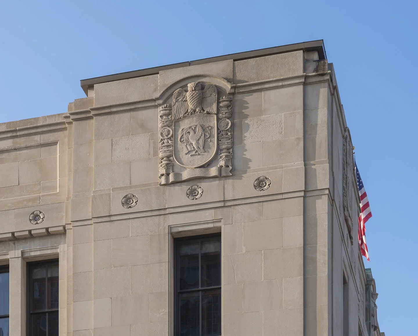 Close-up of a beige stone building with an ornate shield crest featuring an eagle and a star, with decorative columns, and an American flag hanging on the side under a clear blue sky.