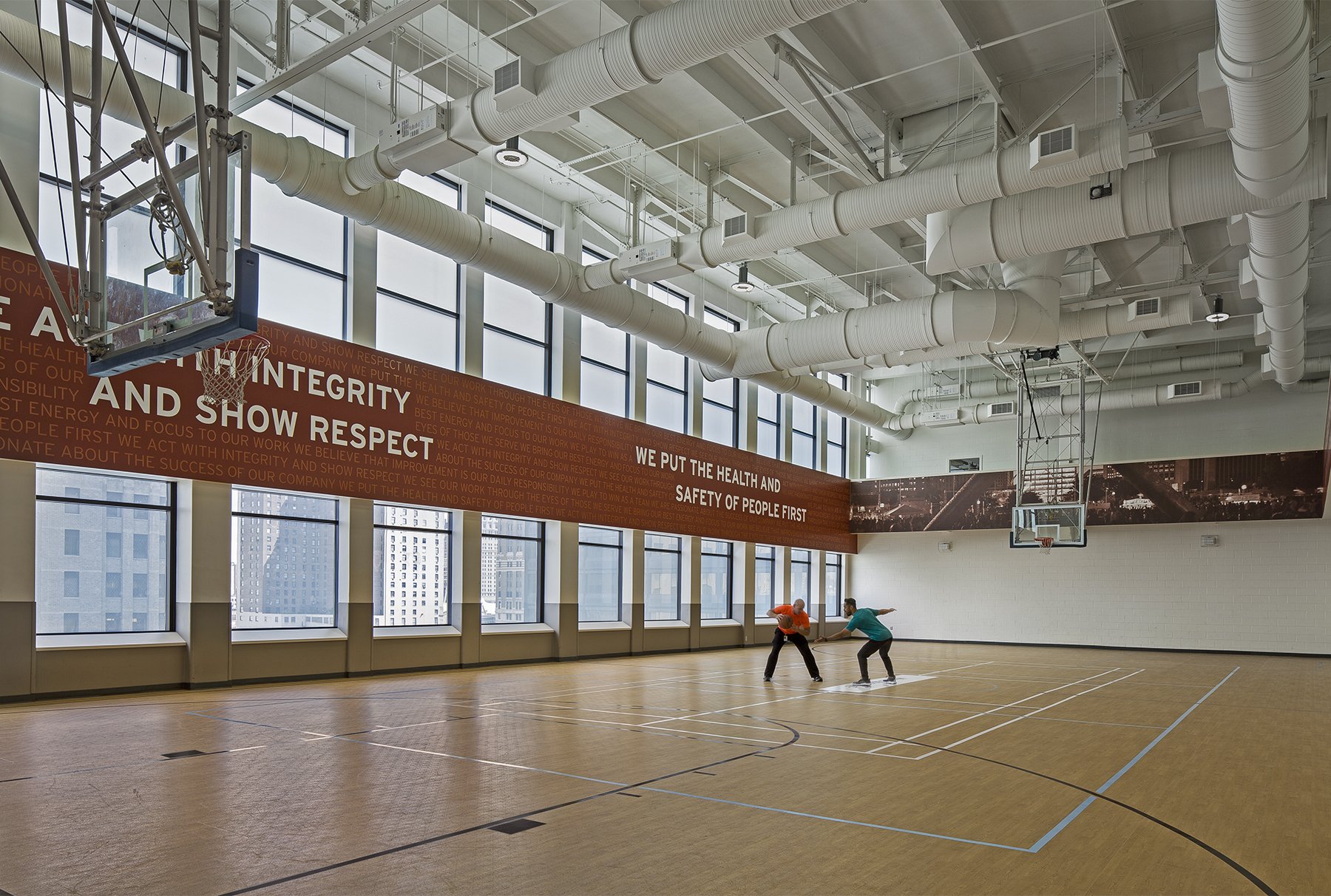 Indoor basketball court with large windows, white ceiling with exposed ducts, and a red banner with white text. Two people are practicing basketball on the court.