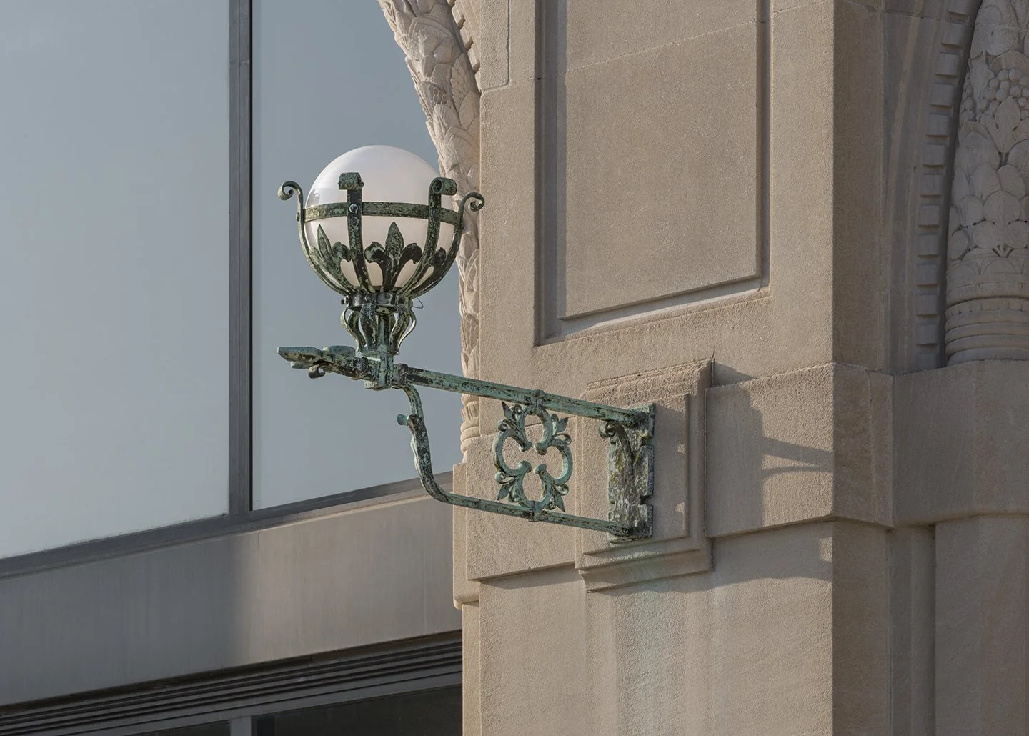 An ornate antique wall-mounted lamp with a glass globe on a decorative metal arm attached to a beige building exterior, with detailed architectural molding near the lamp.