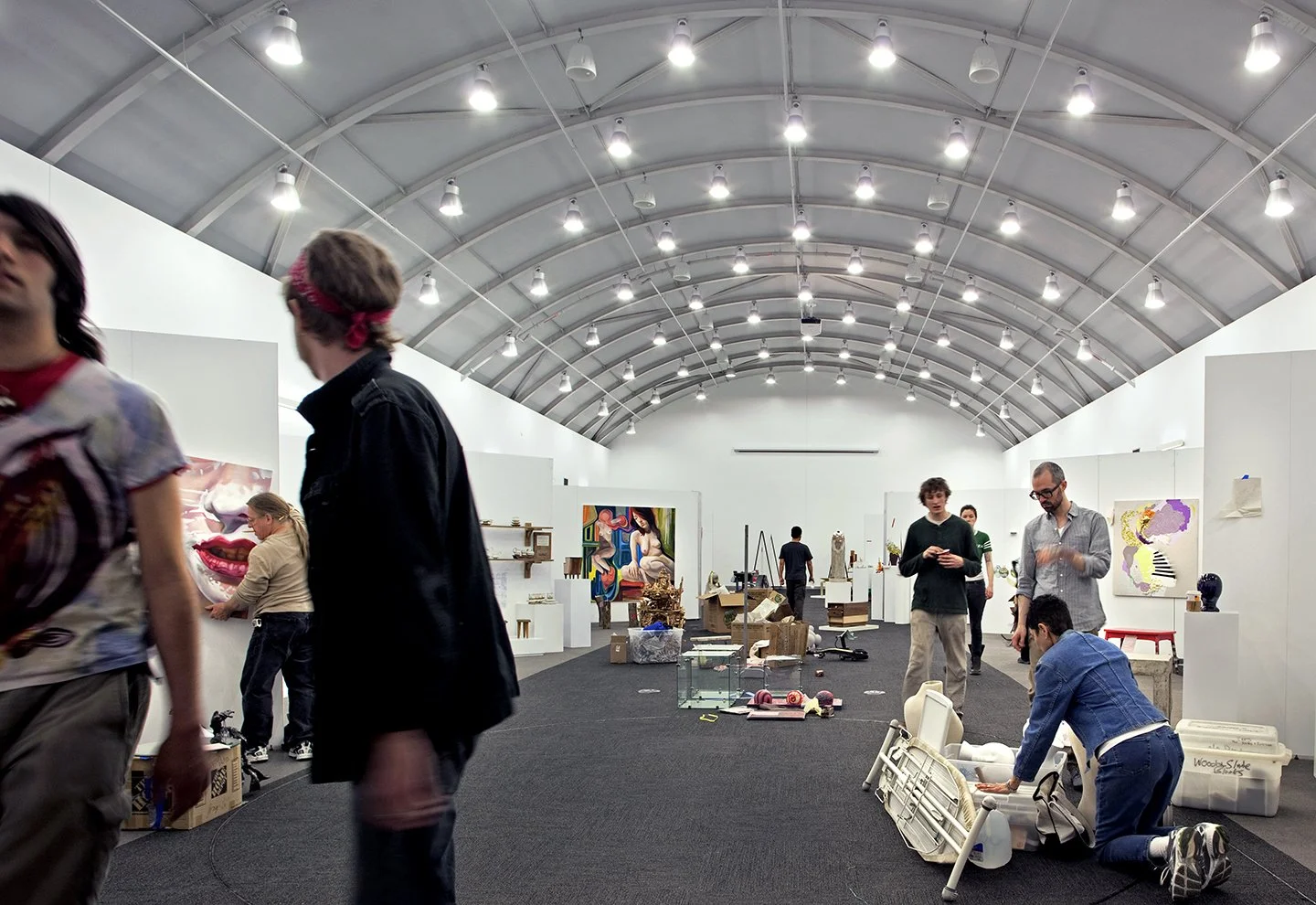 People inspecting artwork and sculptures inside an art exhibition hall with a high curved ceiling and bright lights.