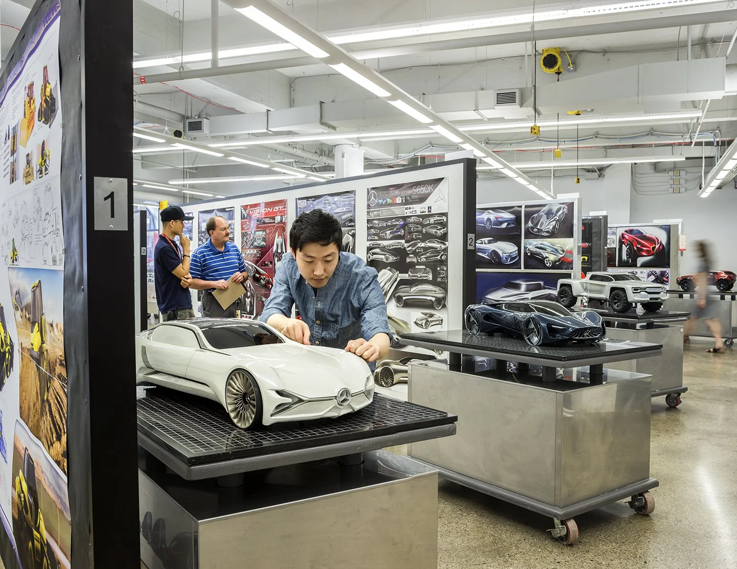 Young man examining a detailed scale model of a luxury MERCEDES-BENZ car on a display table in an automotive design showroom with other car models and design sketches on the walls.
