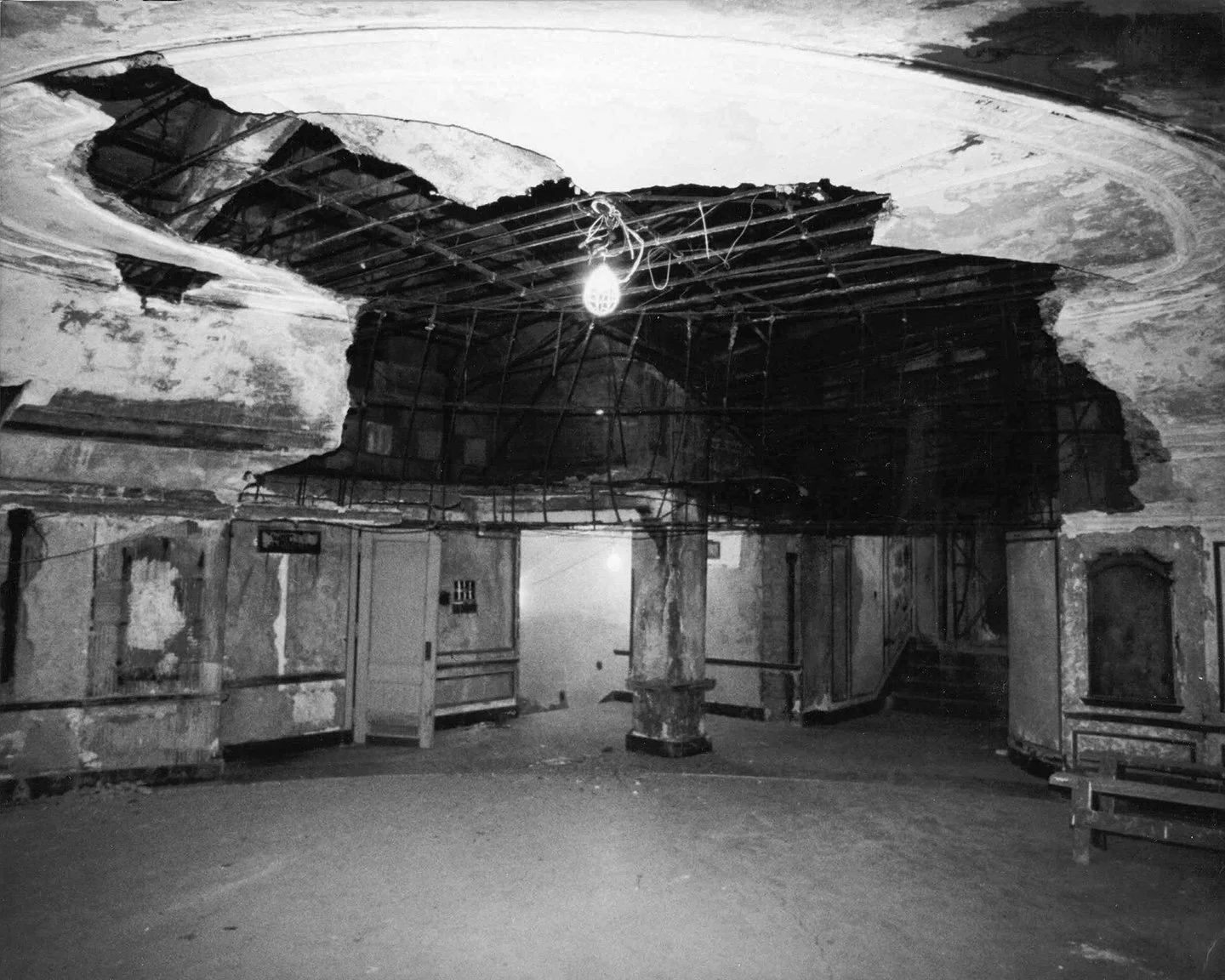 The interior of a building with a damaged ceiling, exposed support beams, peeling paint on the walls, and some doors and furniture. There is a single hanging light bulb.