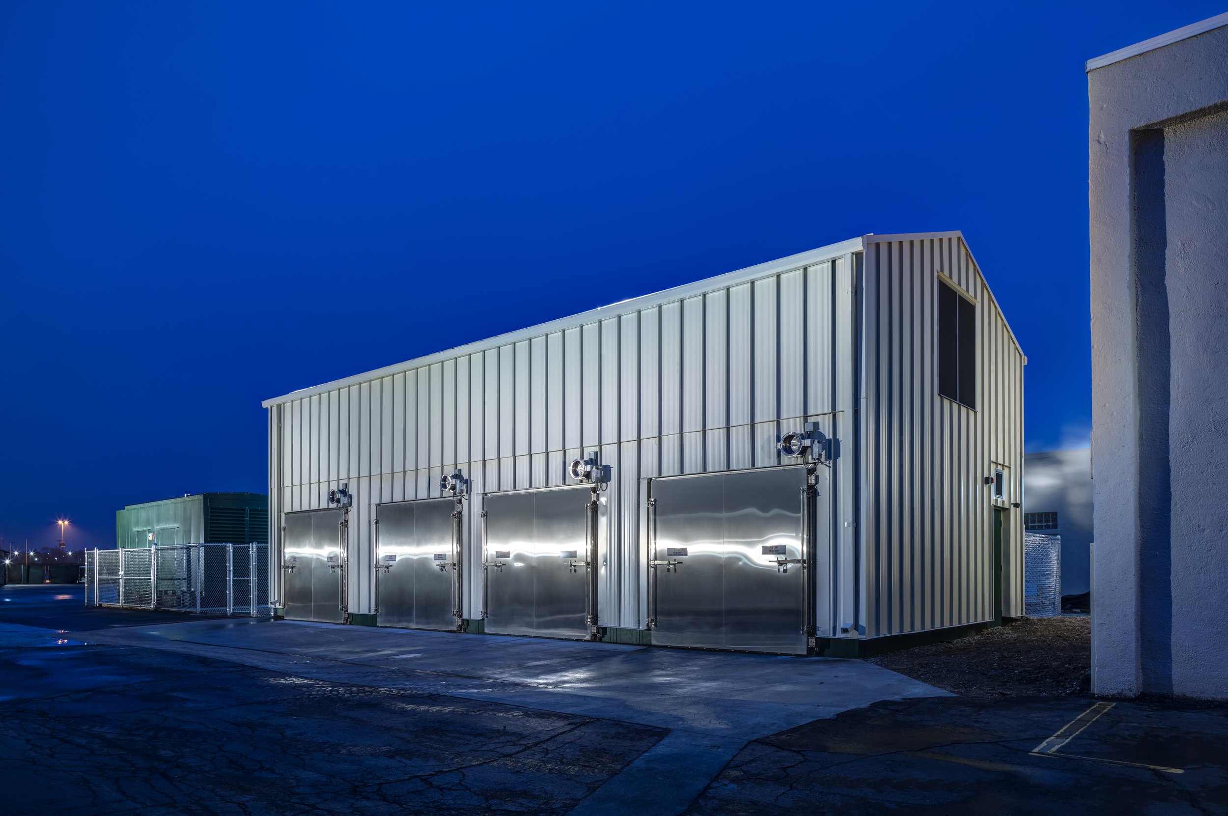 A large industrial building made of metal with several garage doors, illuminated by bright lights at night, with a dark paved parking lot in the foreground and a chain-link fence surrounding the area.