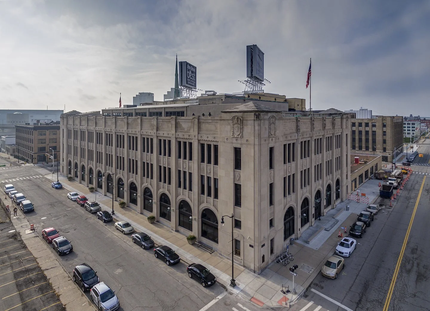 An old multi-story building with arched windows on the ground floor, situated on a city street with parked cars and bike racks. The building has flags on its rooftop and construction work is visible on the surrounding streets.