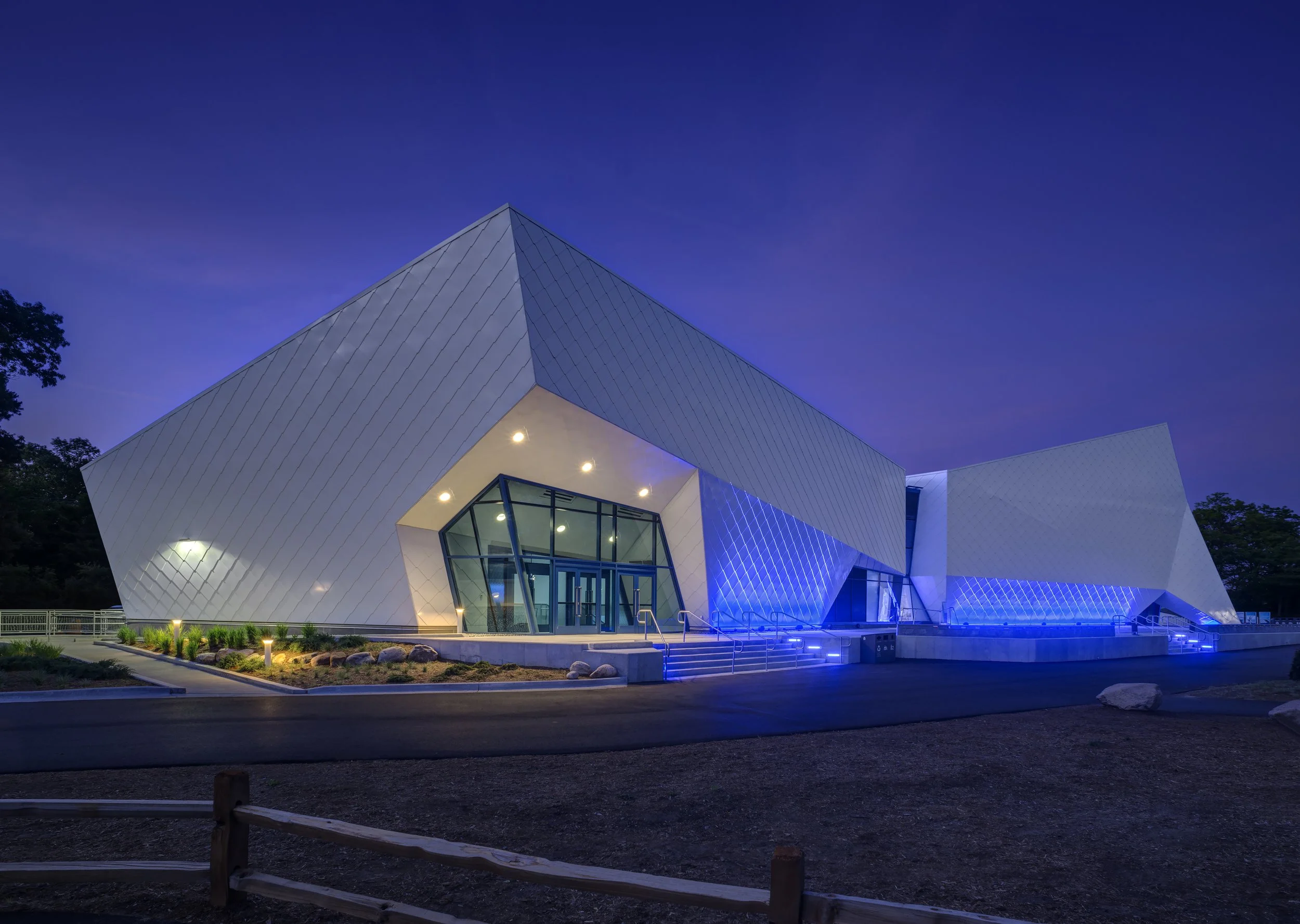 Modern white building with angular design, large glass windows, and blue accent lighting at night, surrounded by trees and a manicured landscape.
