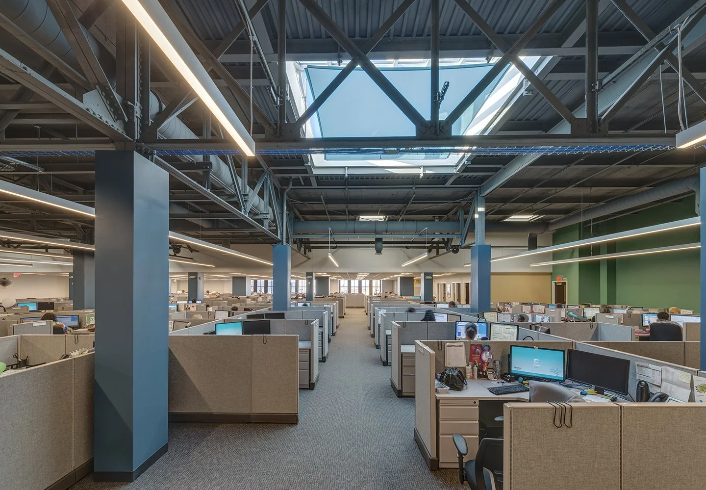 Office cubicles in a modern, open-layout workspace with high ceilings and skylights, featuring several employees working at their computers.