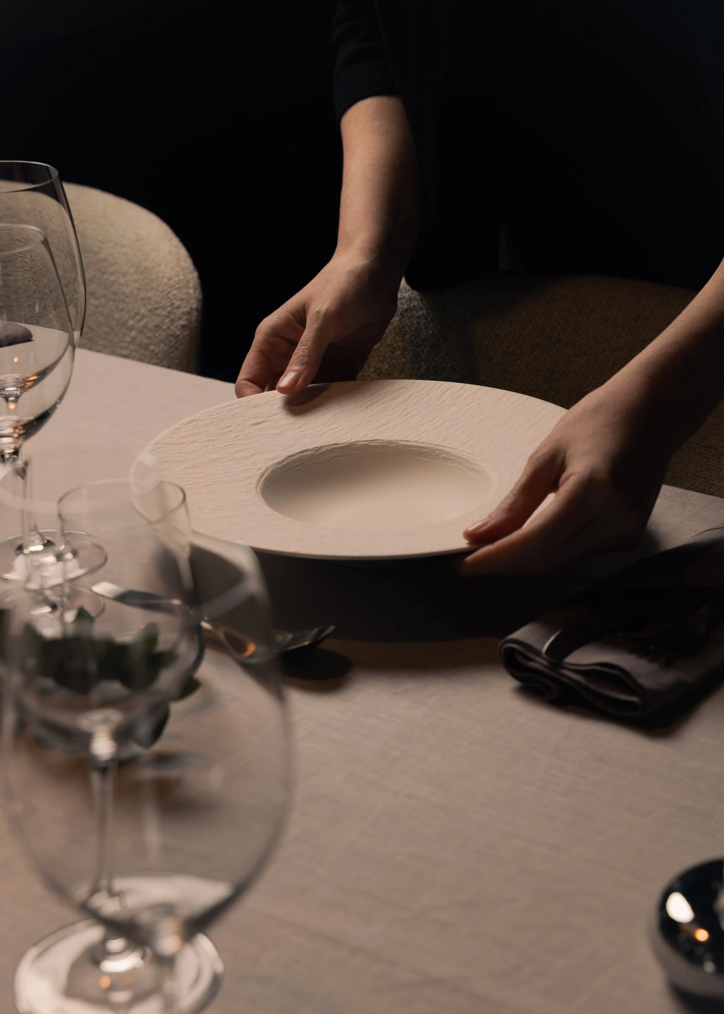 Person placing a beige plate with a textured surface on a dining table set with wine glasses, silverware, and a folded napkin.