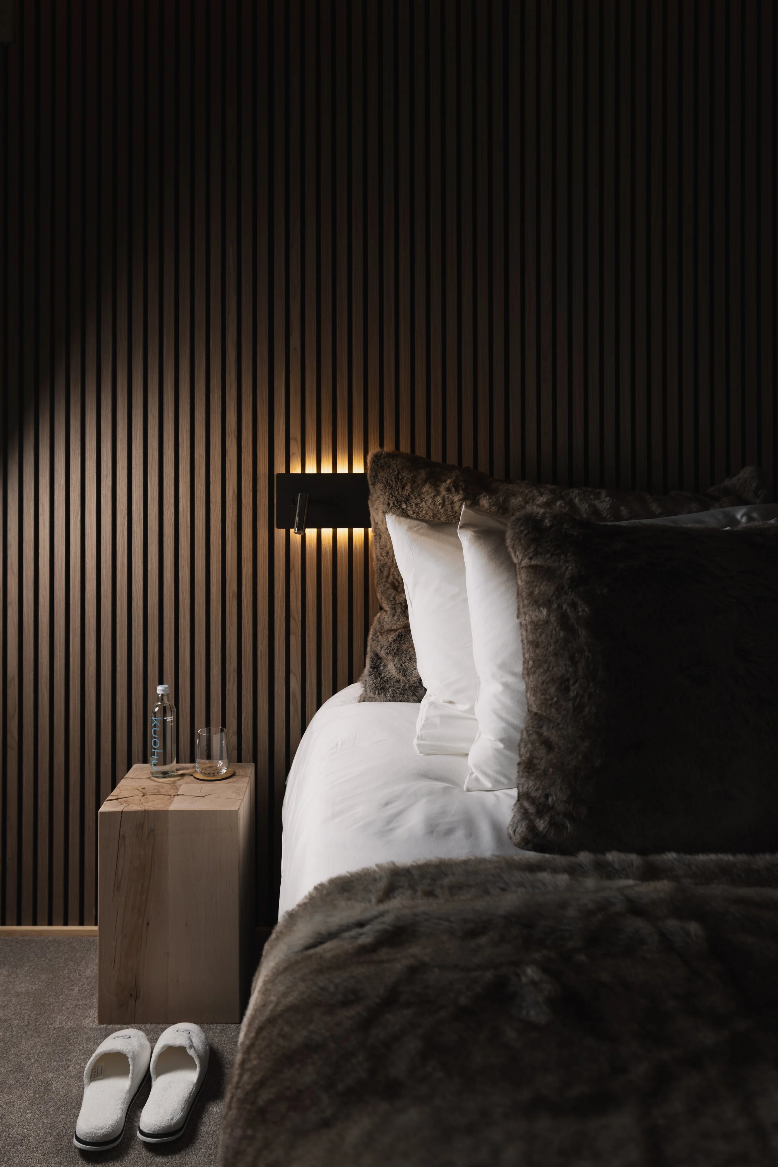 A bedroom with dark wooden slat wall, a side table with water bottle and glass, white slippers on carpet, and bed with white bedding and dark fur throw pillows.