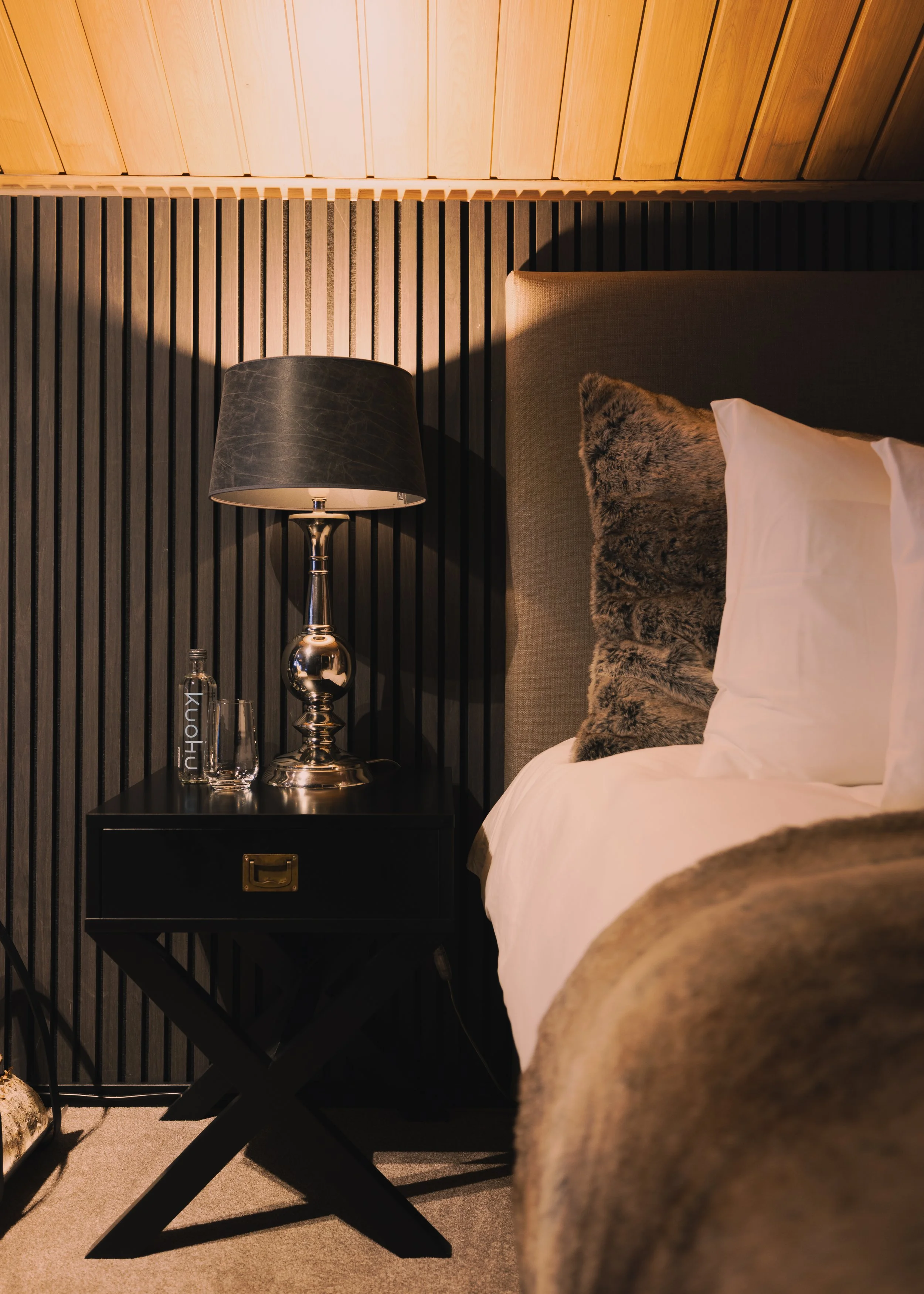 Close-up of a modern bedroom corner with a bedside table, a lamp, and pillows on the bed, with a textured wall and wooden ceiling.