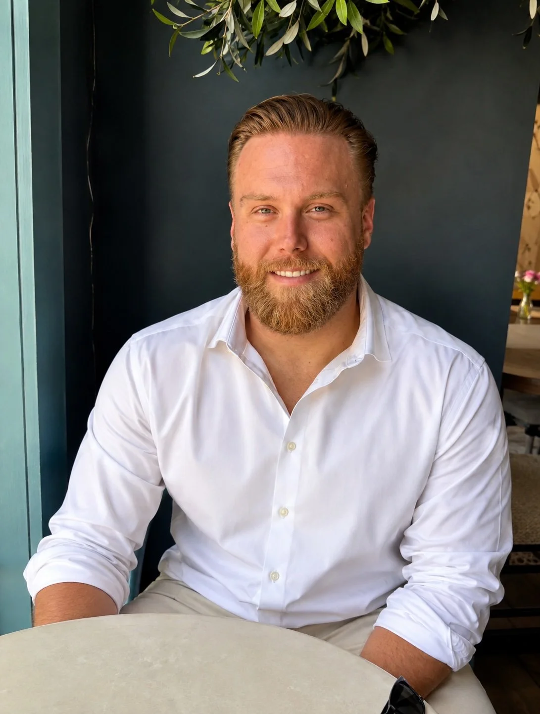 A man with a beard and blonde hair smiling at the camera, wearing a white shirt, sitting at a table in a setting with dark walls, green plants overhead, and a vase with flowers in the background.