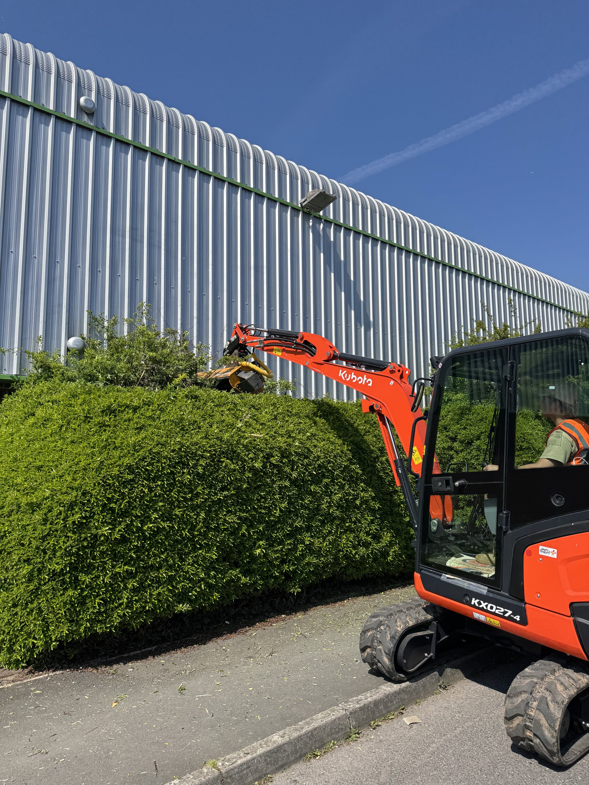 Kubota 2.5 ton digger with flail trimming a large green hedge outside a metal building under a clear blue sky.