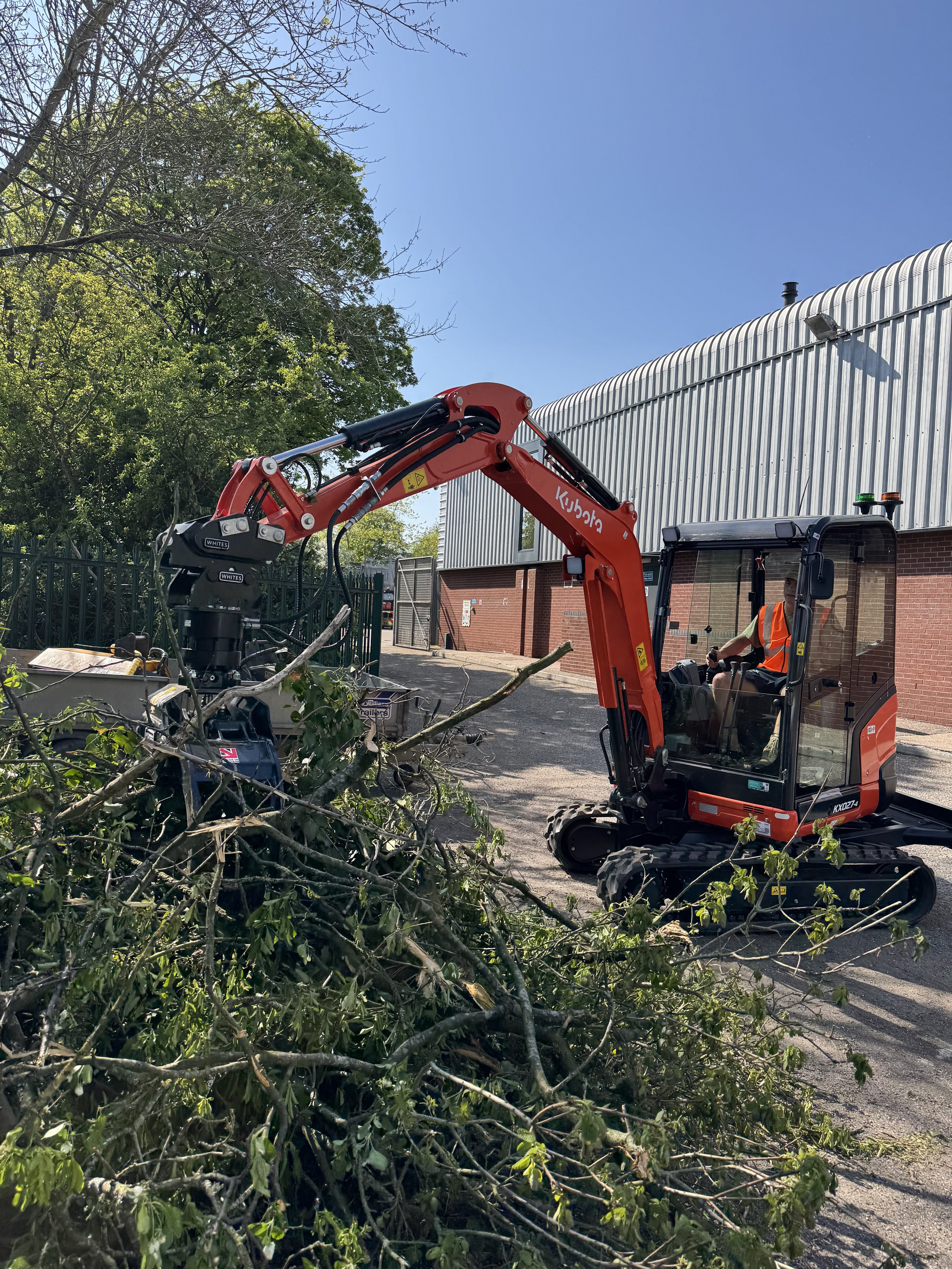 A person operates a Kubota mini excavator with a grapple attachment, clearing fallen tree branches in an outdoor area near a metal-sided building on a sunny day.