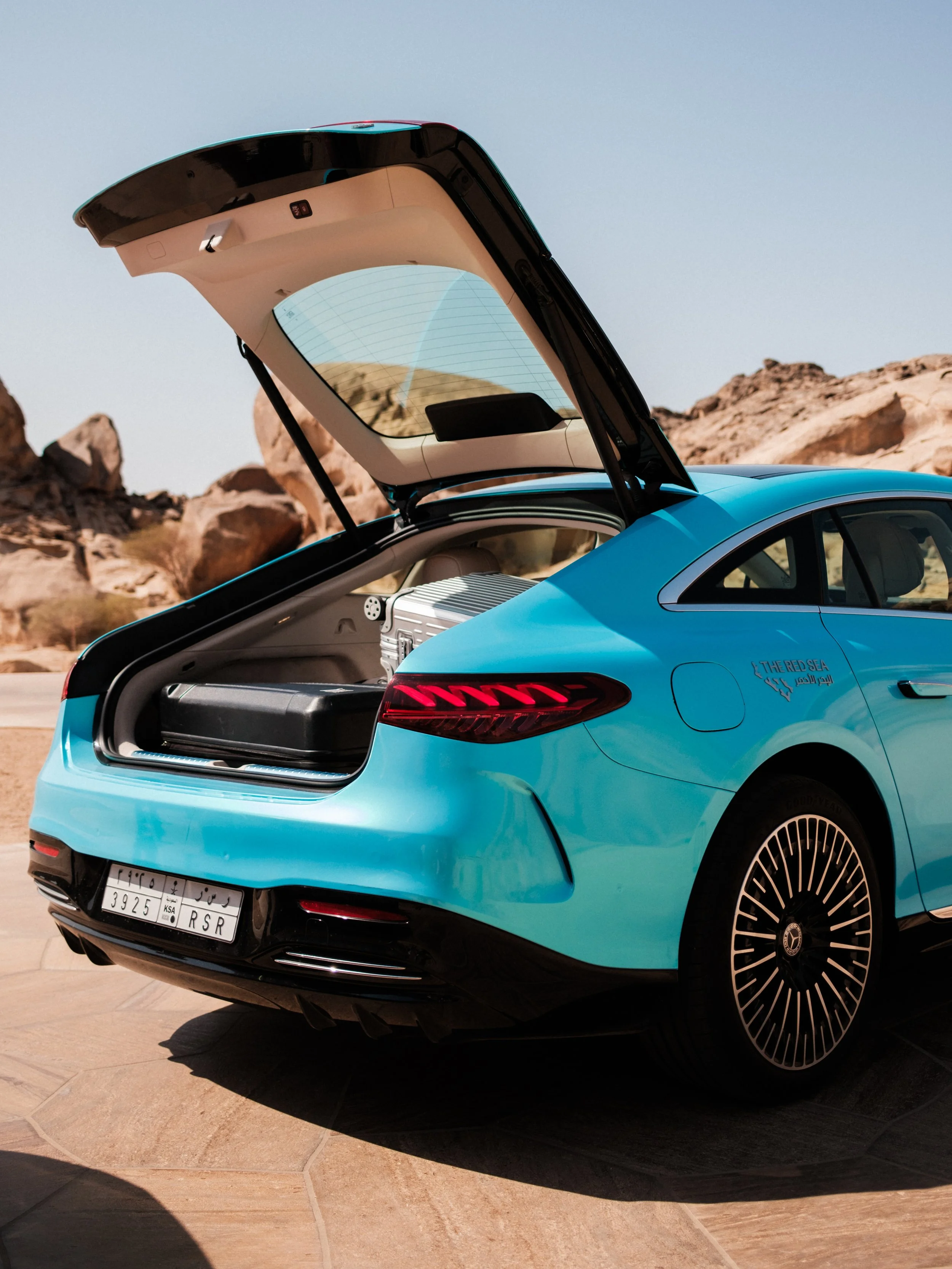 A woman sitting on a lifeguard stand on a beach, taking a photo of a man in front of her with his back to the camera. The woman wears a green and white dress, and the man is dressed in white. The beach has white sand and the ocean in the background with a clear blue sky.
