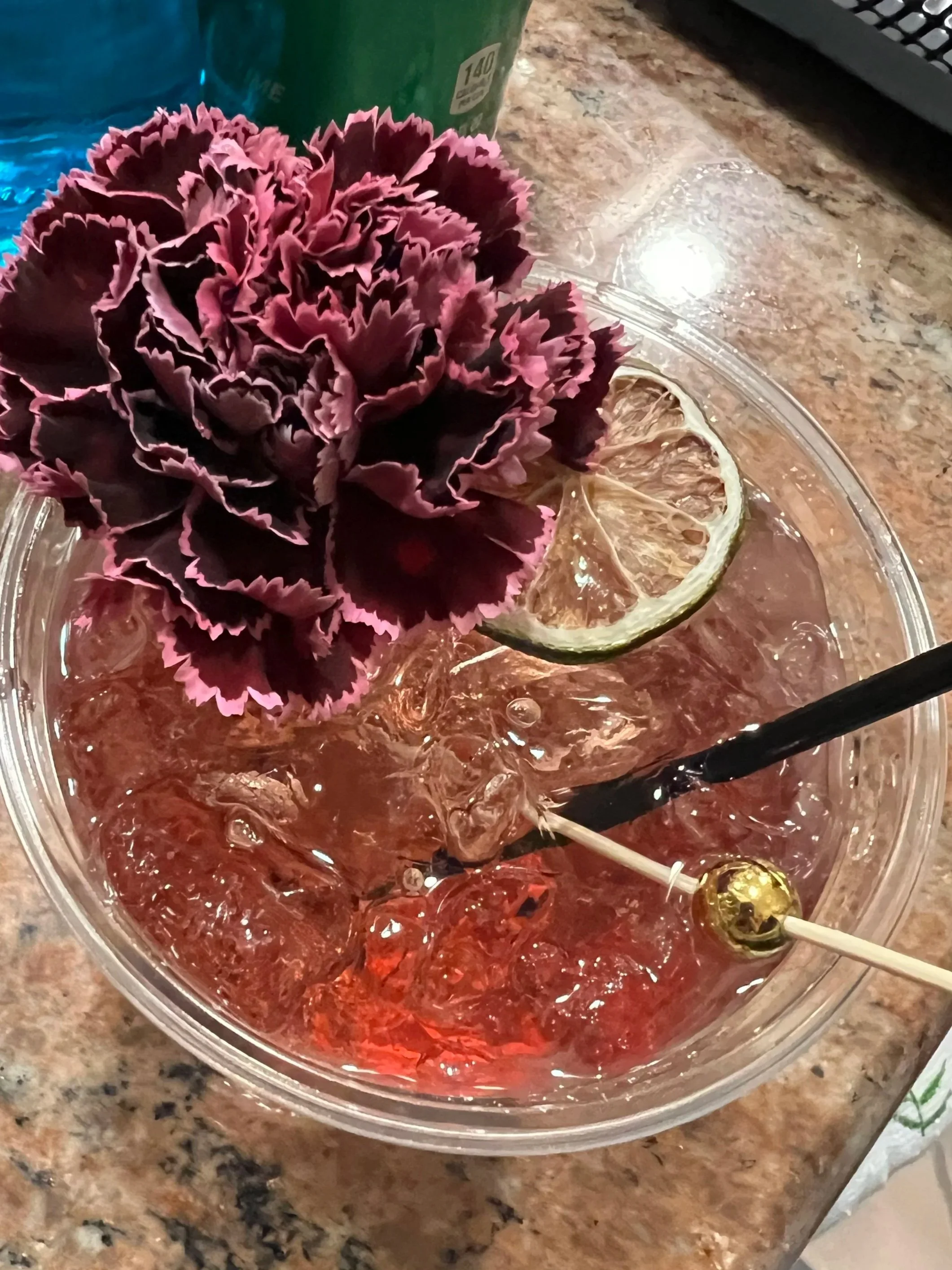 A pink and purple decorative flower garnishing a cocktail with a lemon slice and ice in a clear glass.