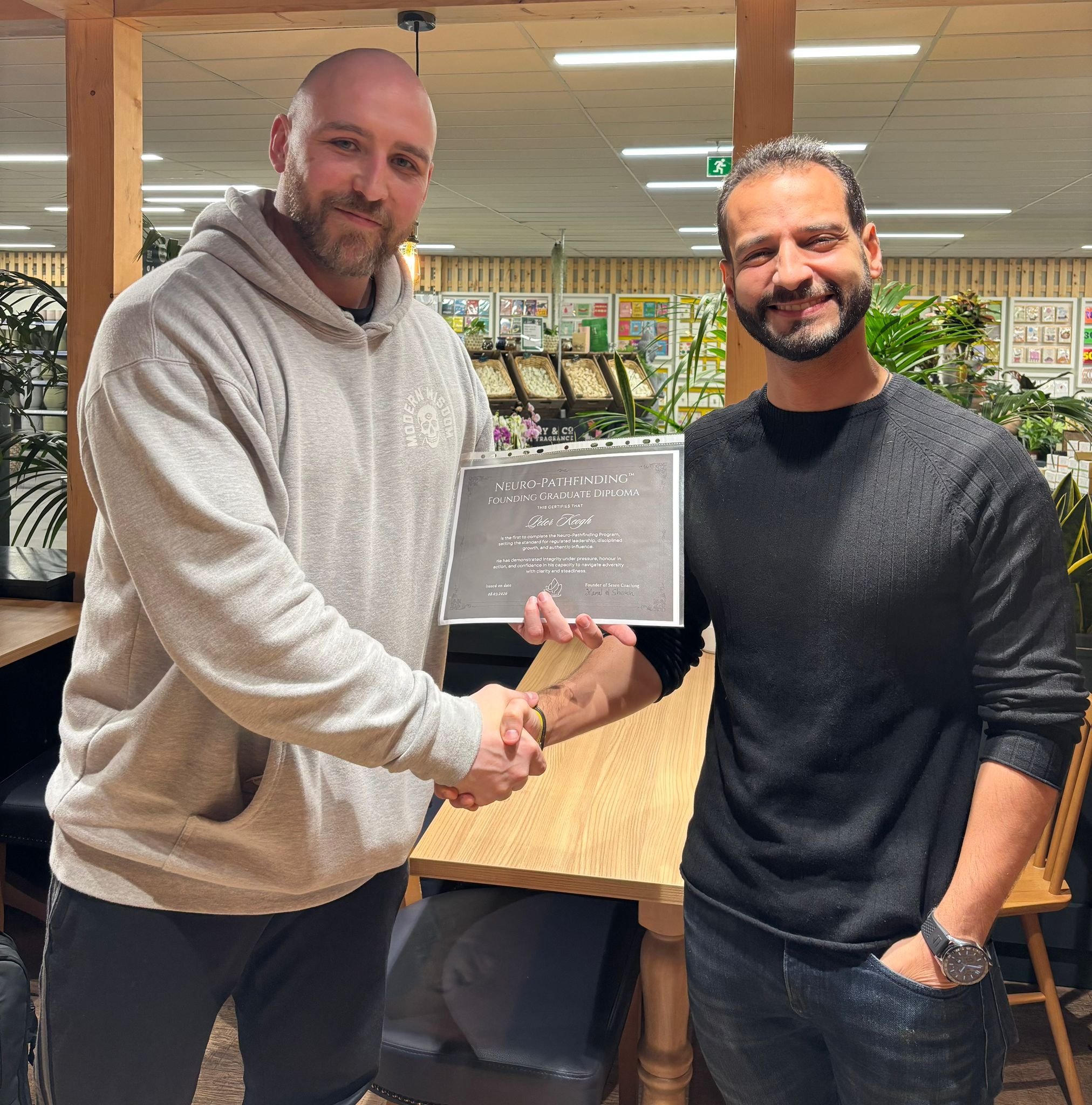 Two men shaking hands indoors, with one holding a diploma. The background features shelves with books and plants, and a wooden table.