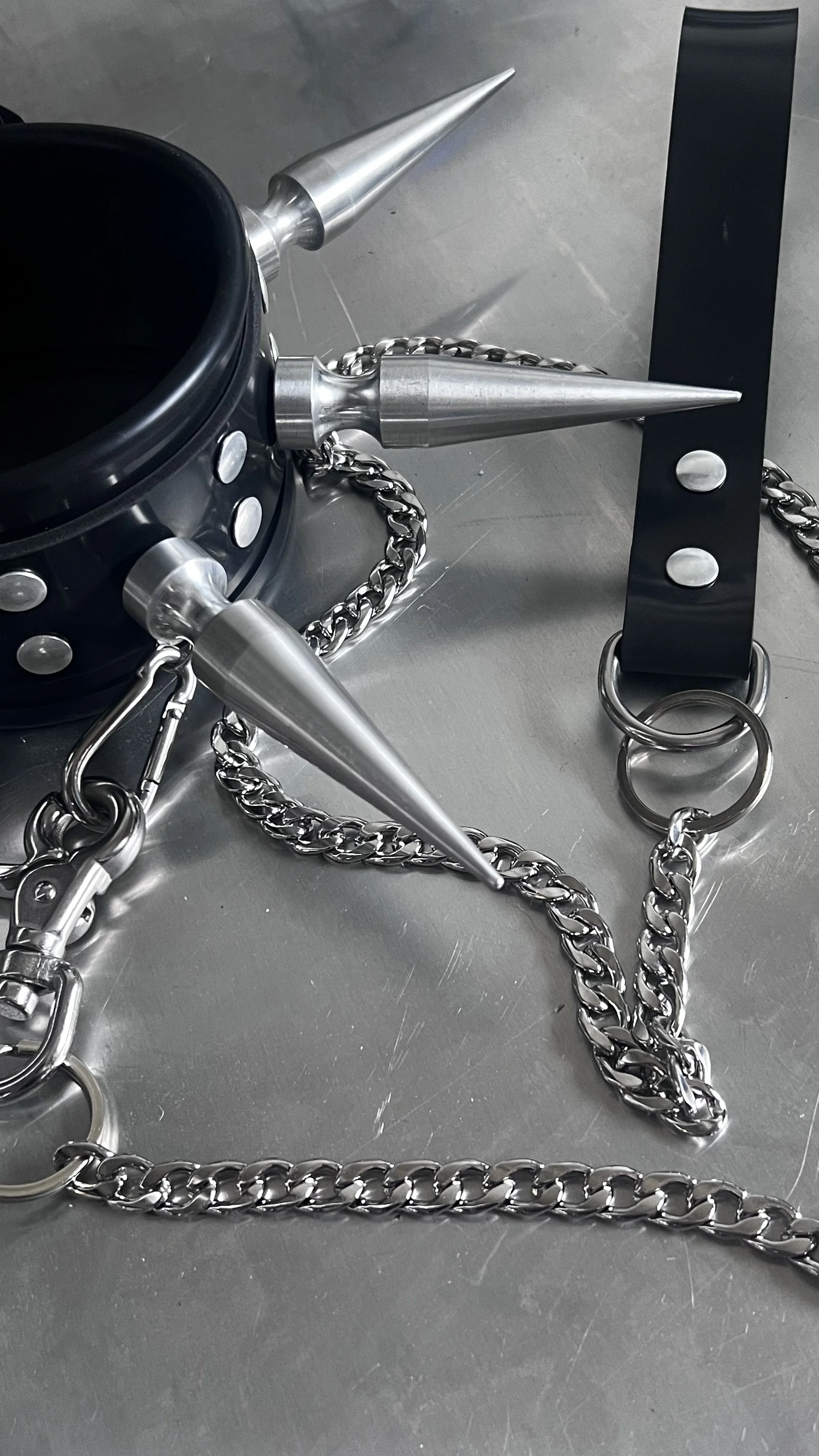 Close-up of a metallic chain, several pointed metallic cones, and a black belt with silver buttons on a gray surface.