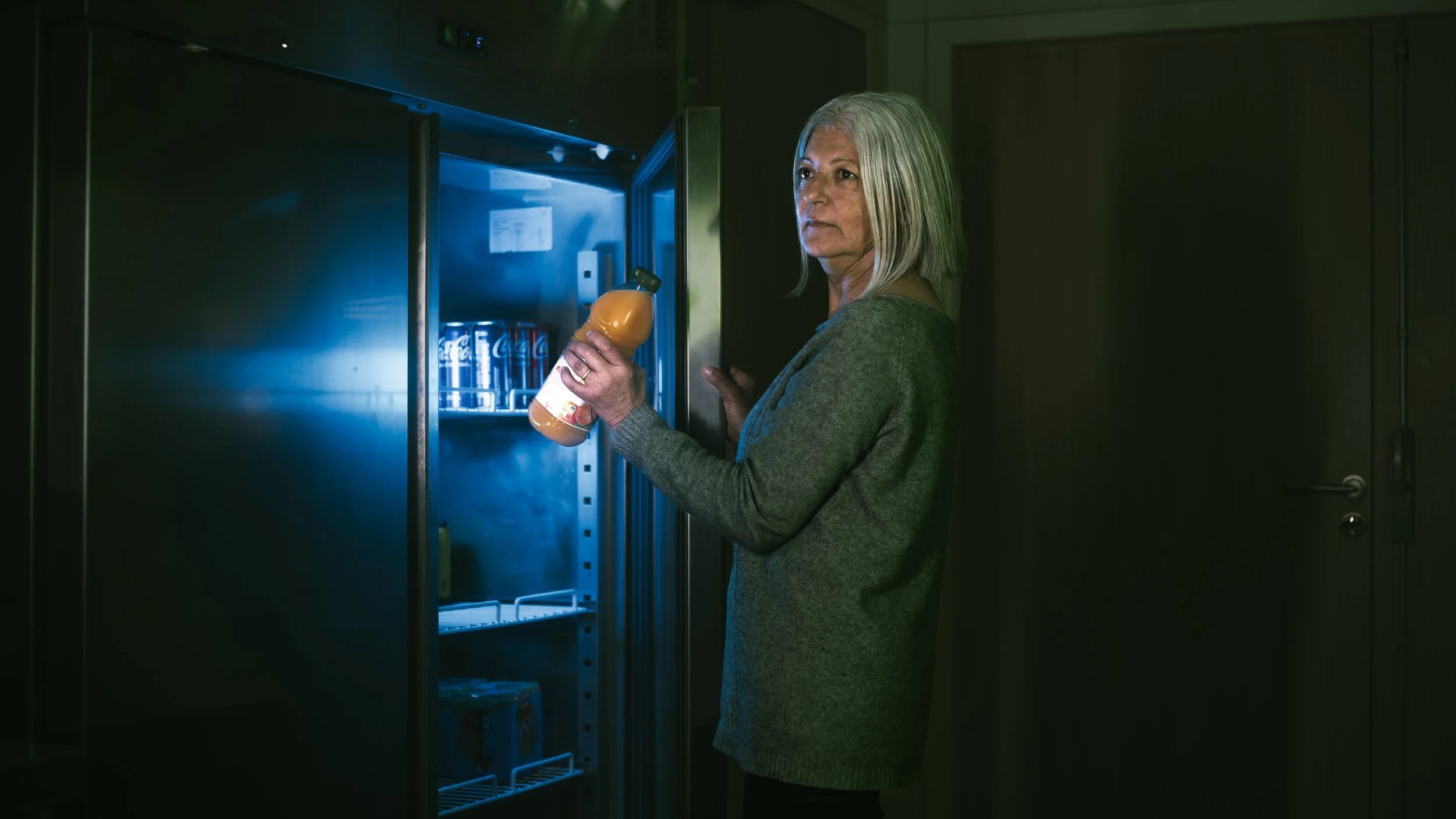 Une femme âgée avec des cheveux gris clair, portant un pull gris, se tient devant un réfrigérateur et tient une bouteille de jus d'orange dans la main, dans une pièce sombre.